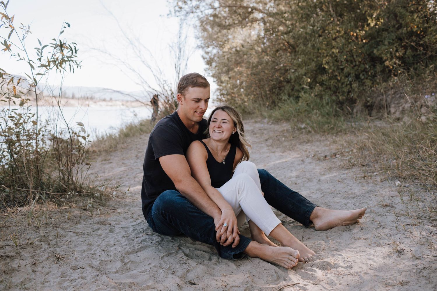 A couple relaxes together on a sandy beach trail with water and foliage in the background.