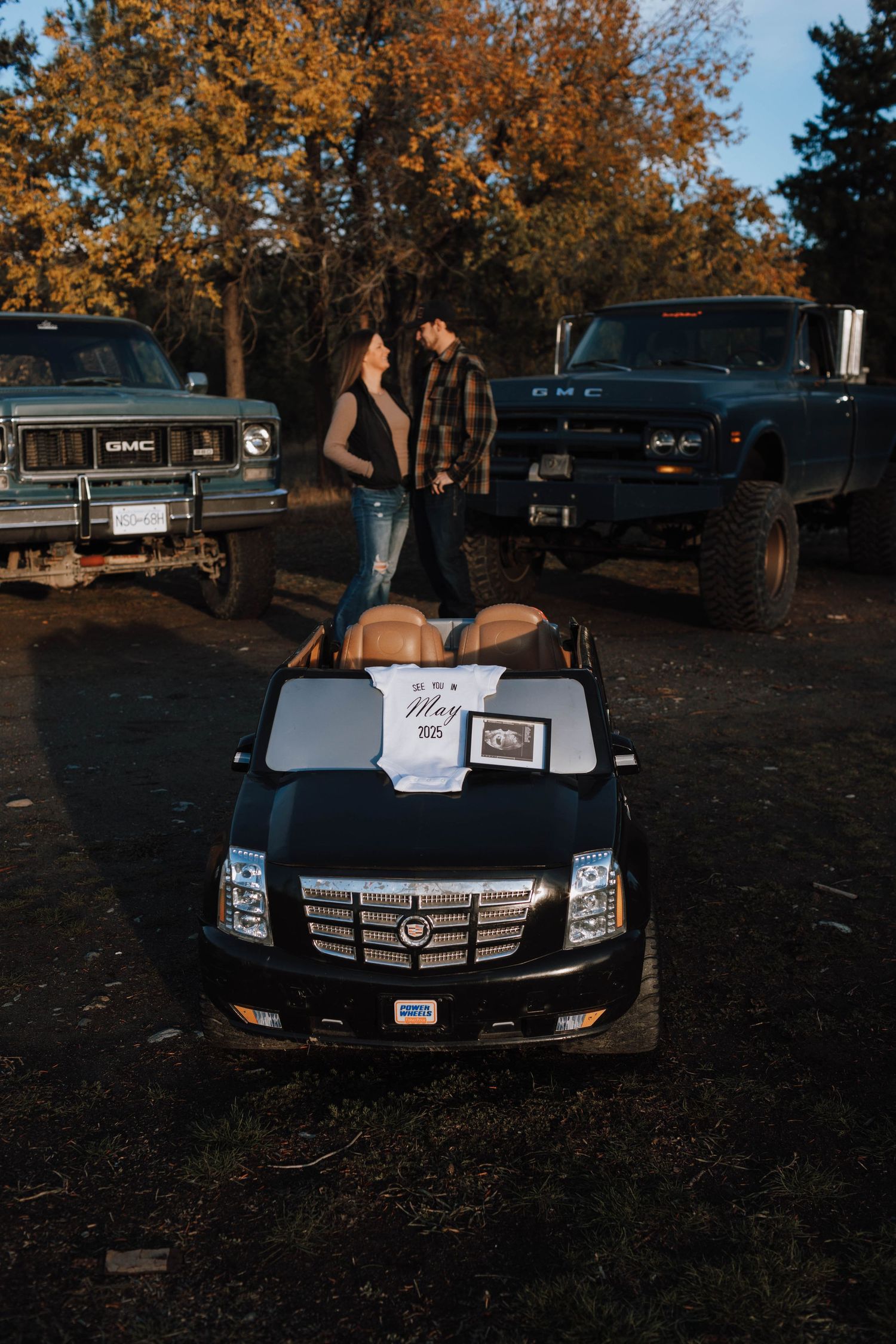 A couple poses with their toy Cadillac Escalade between two large trucks during fall.