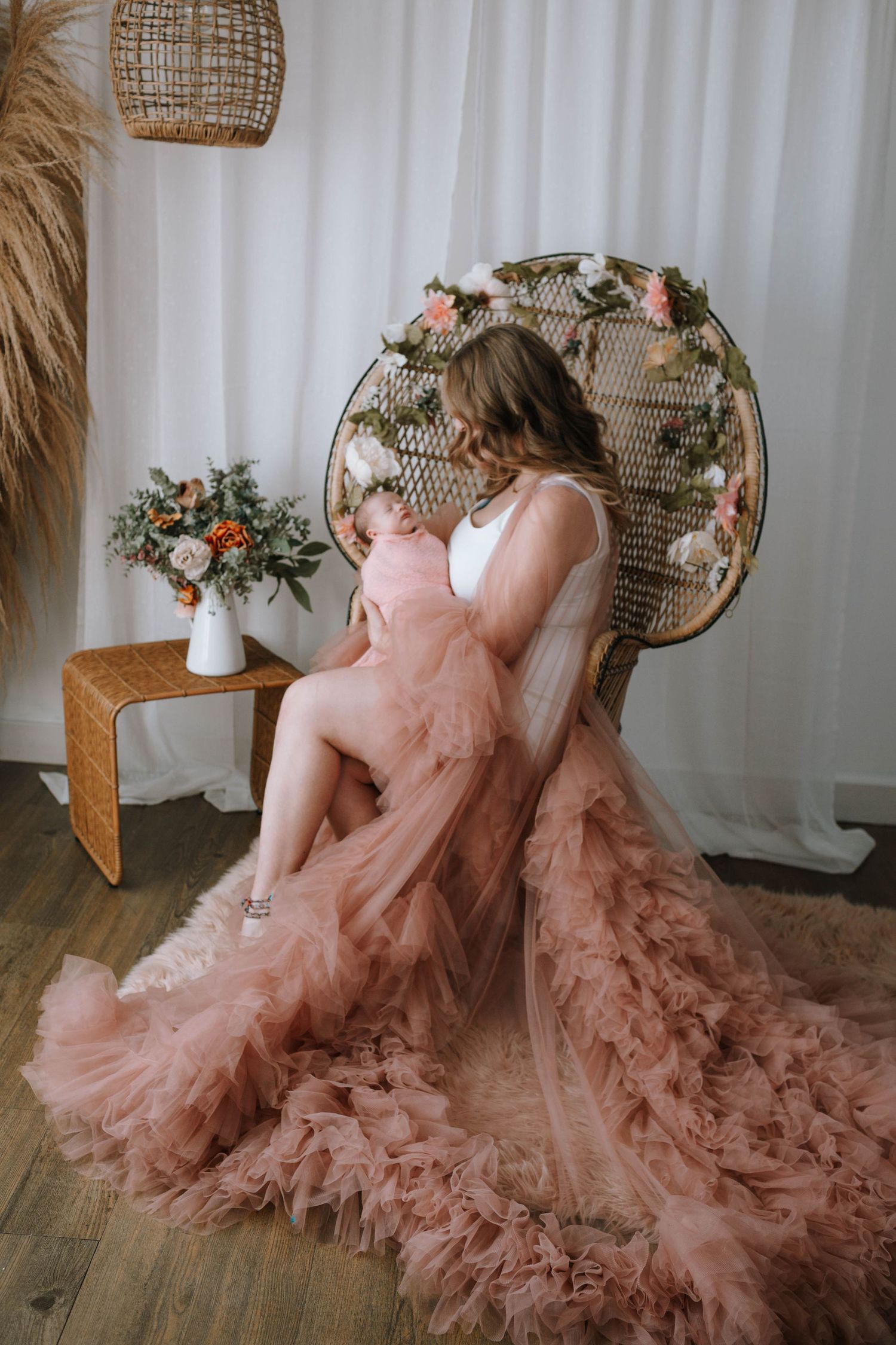 Maternity photo with flowing pink tulle dress and peacock chair surrounded by flowers.