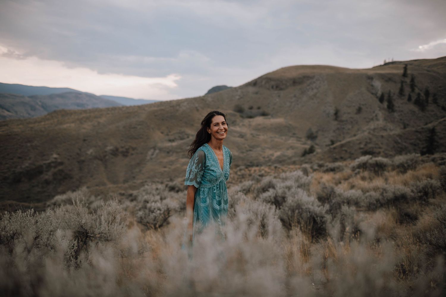 Figure in a teal dress standing among desert brush with mountains in the background.