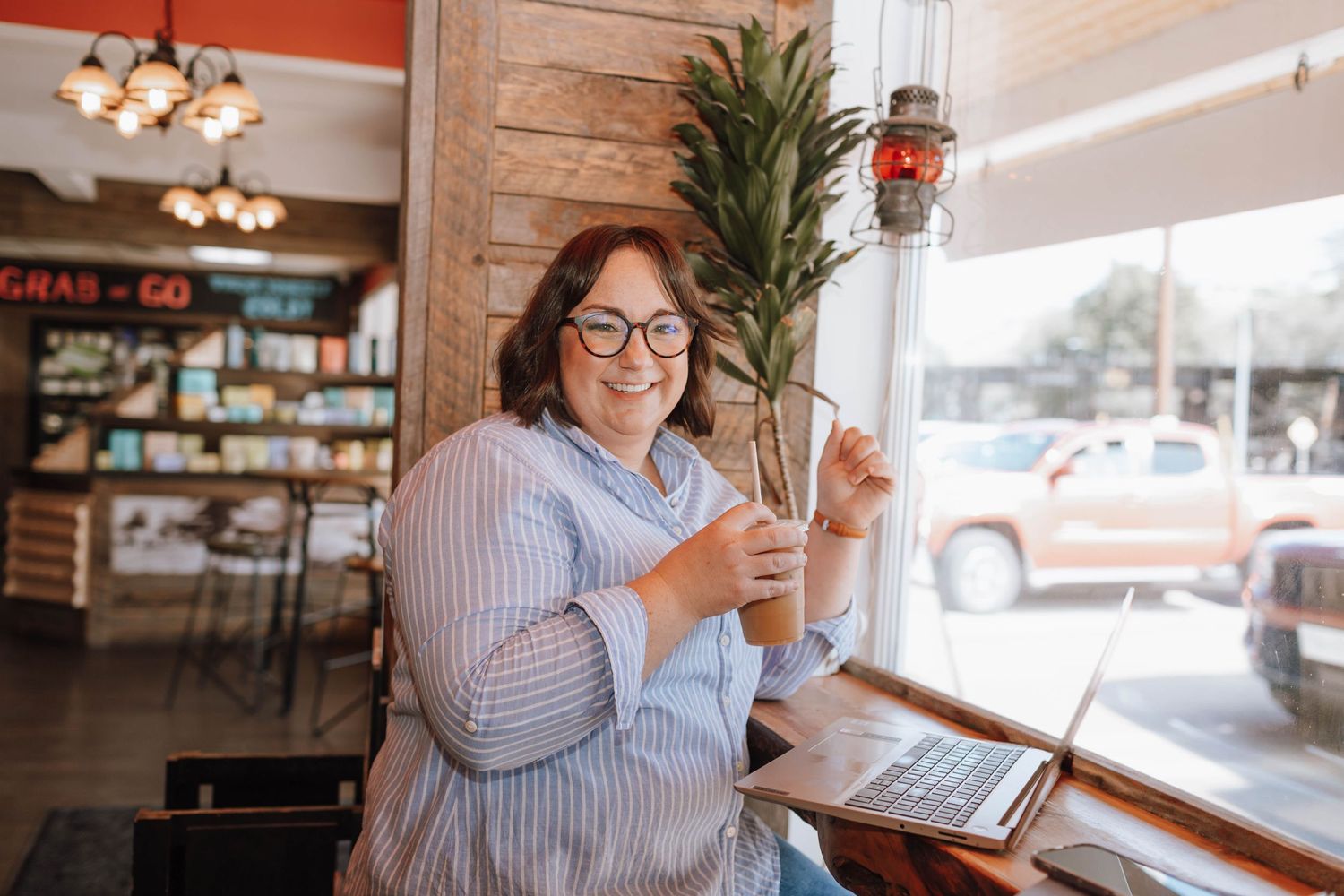 Bright cafe interior with rustic wood walls and large windows showing someone using a laptop.