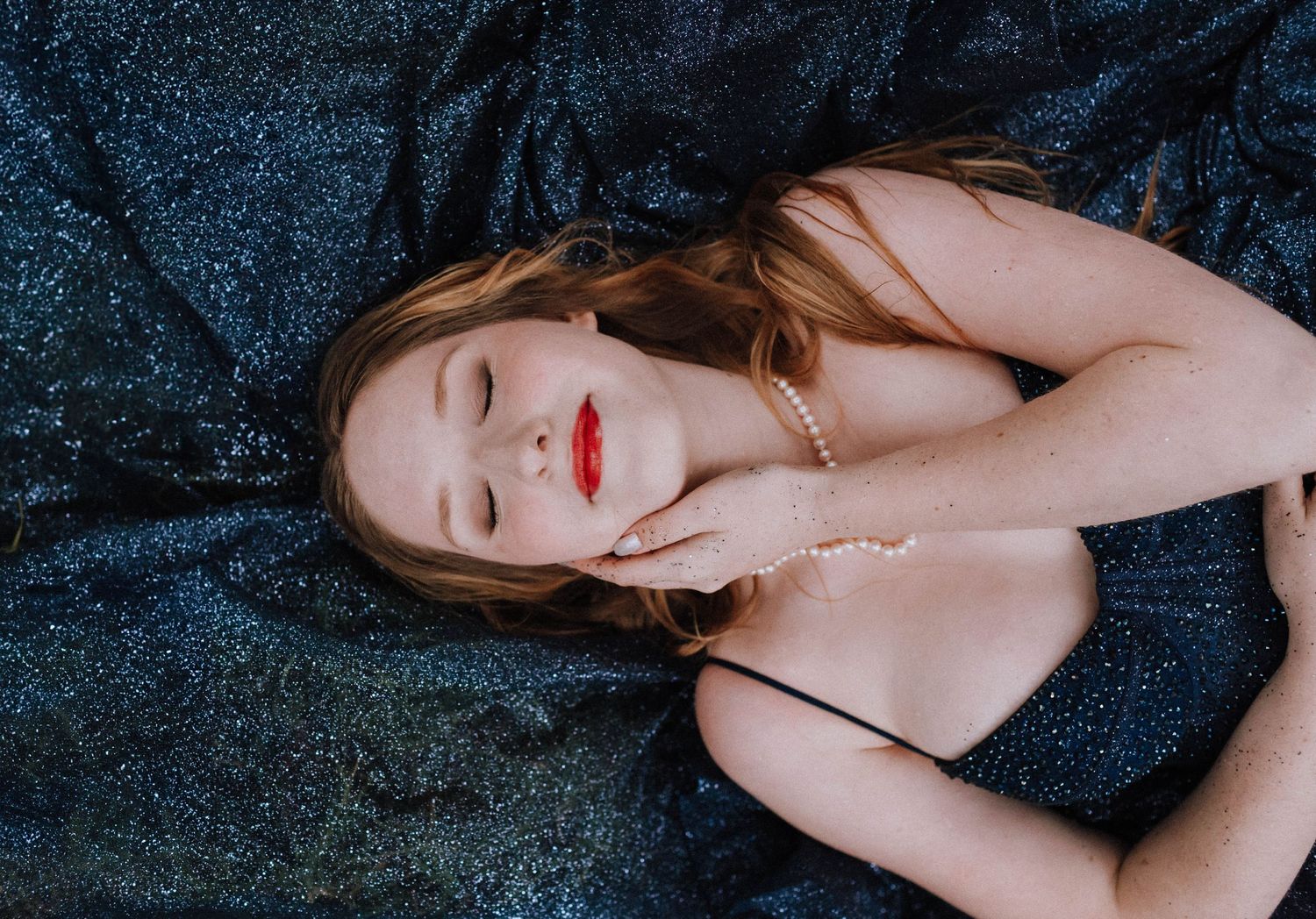Artistic portrait of someone lying on dark blue glittery fabric with red lipstick.