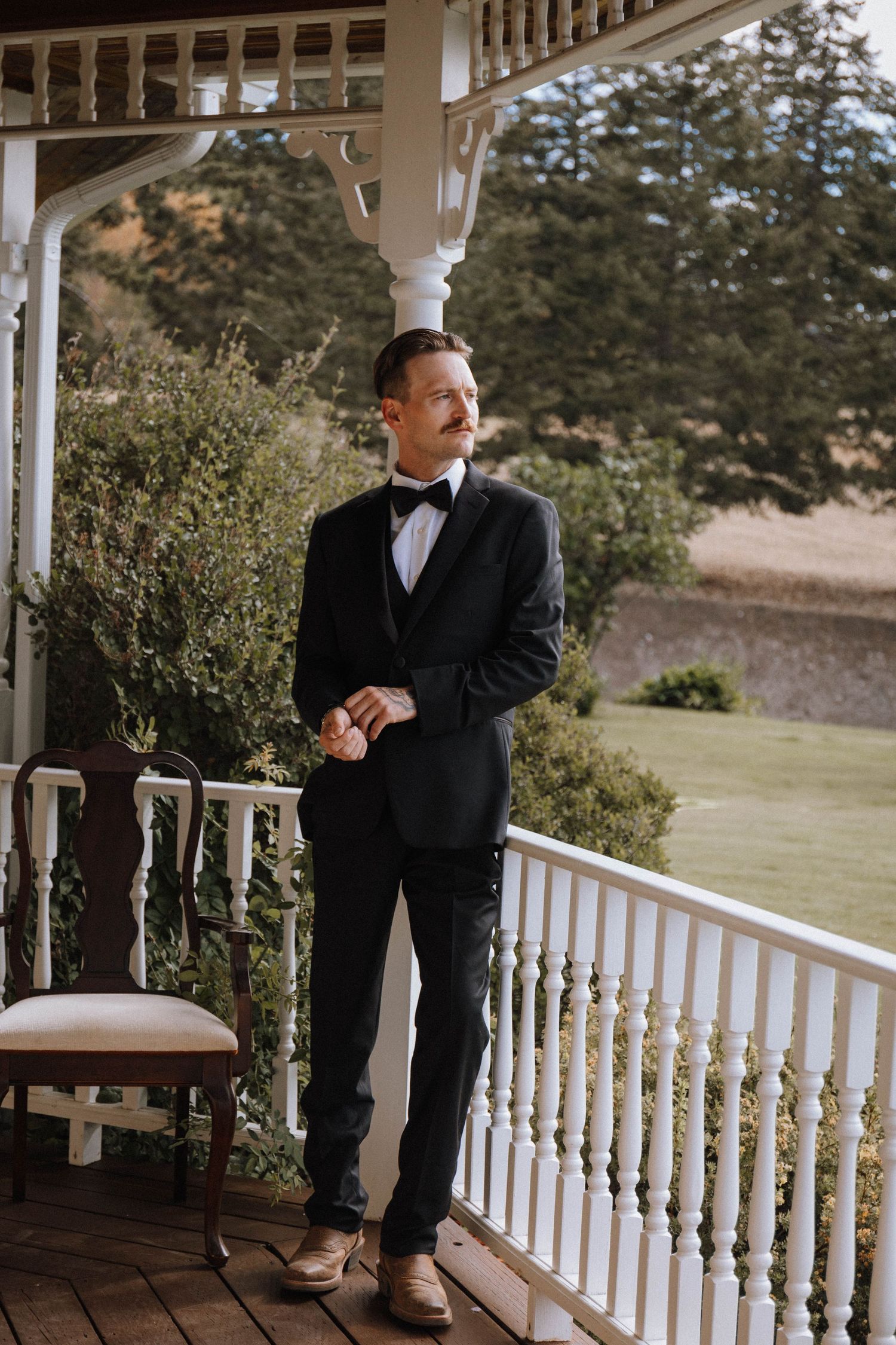 A person in a classic black tuxedo and bow tie stands on an elegant white porch with ornate railings and garden views.