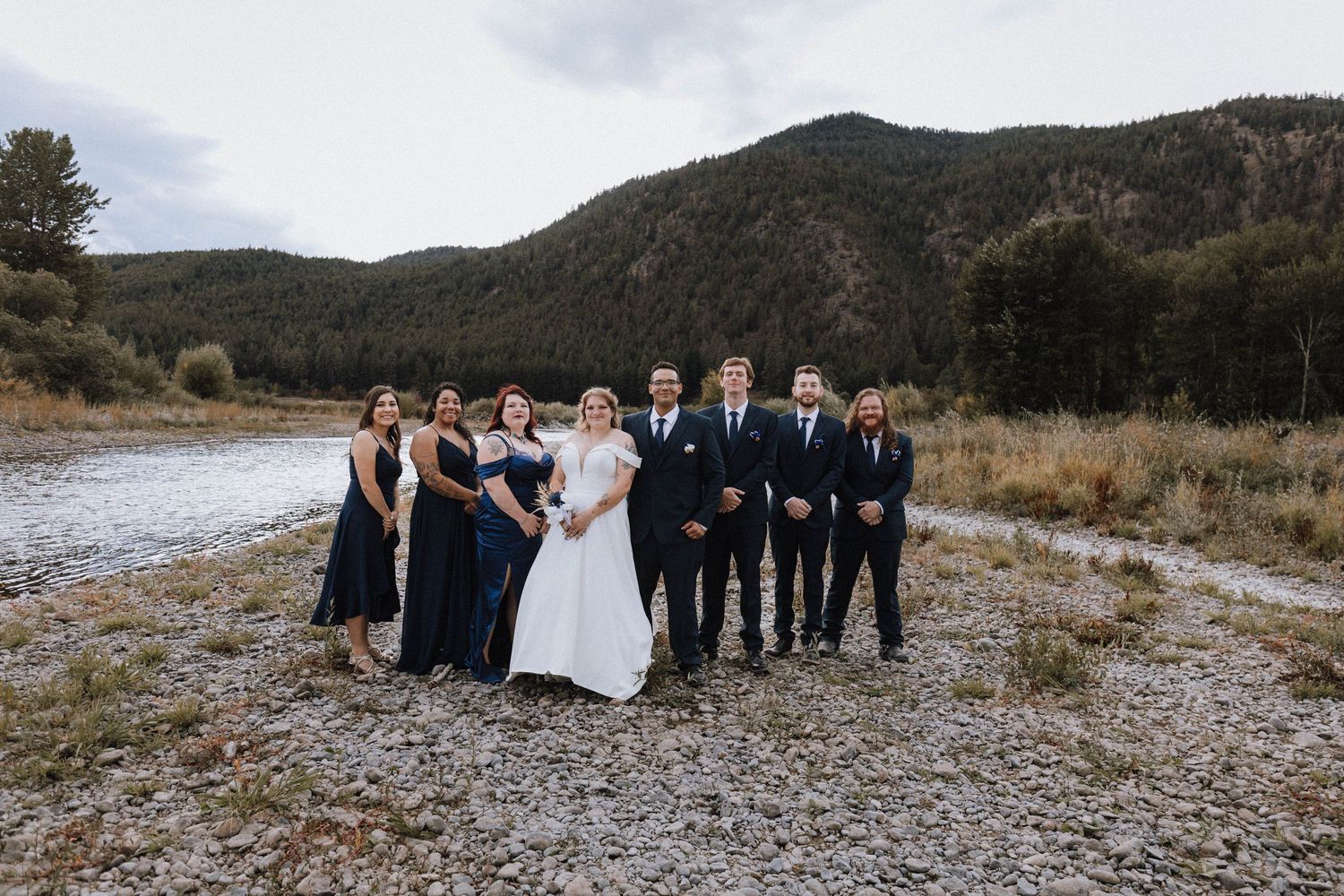 A wedding party stands together on a rocky riverbank with mountains and forest in the background.