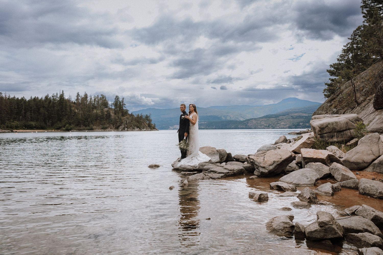 Bride and groom standing on rocky shoreline of a mountain lake under dramatic cloudy skies.