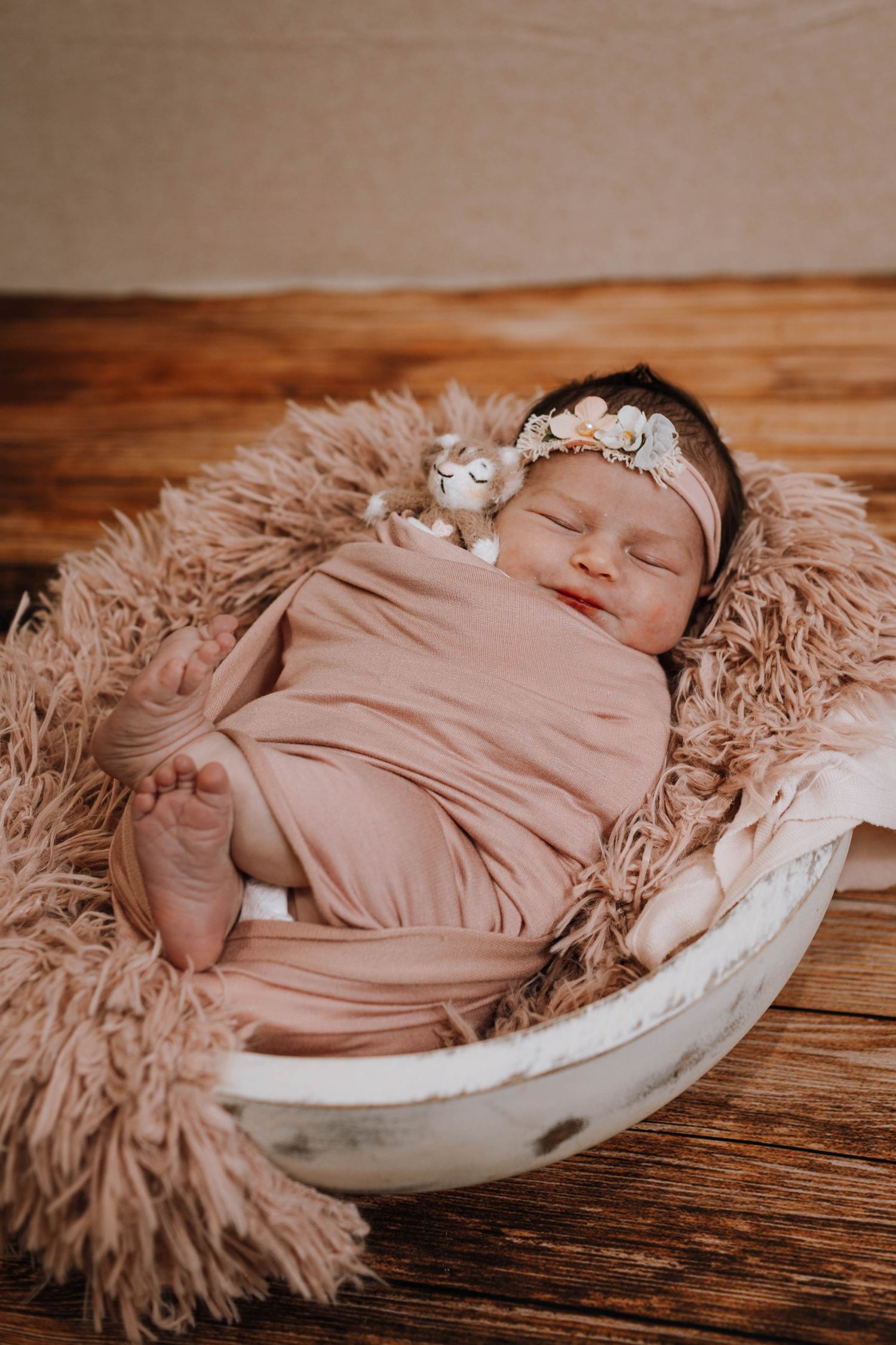 Sleeping newborn wrapped in pink swaddle rests on fluffy beige fur in rustic white bowl with floral headband.