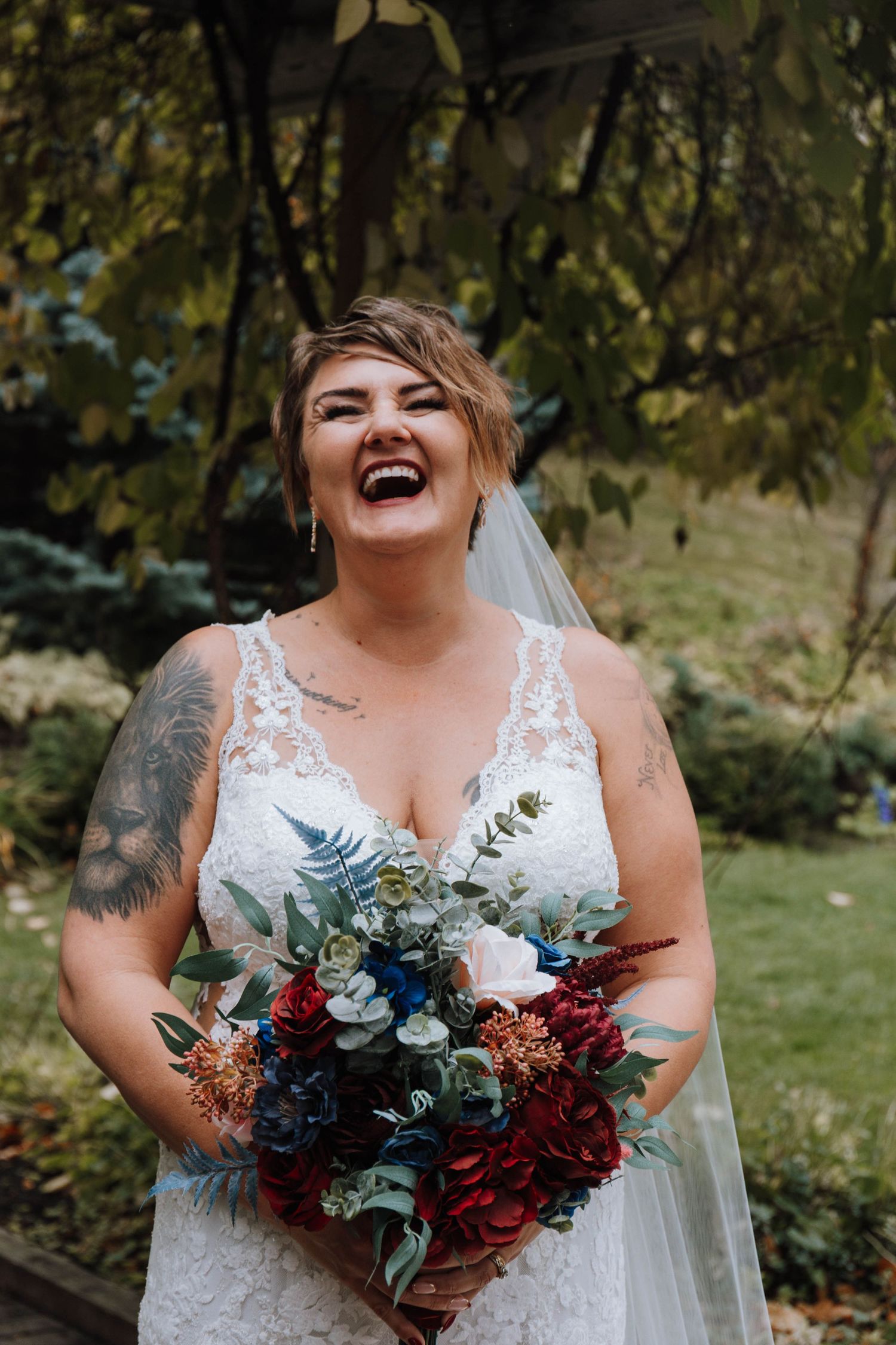 A bride in a lace dress holds a vibrant red and blue bouquet while laughing joyfully in an outdoor garden setting.