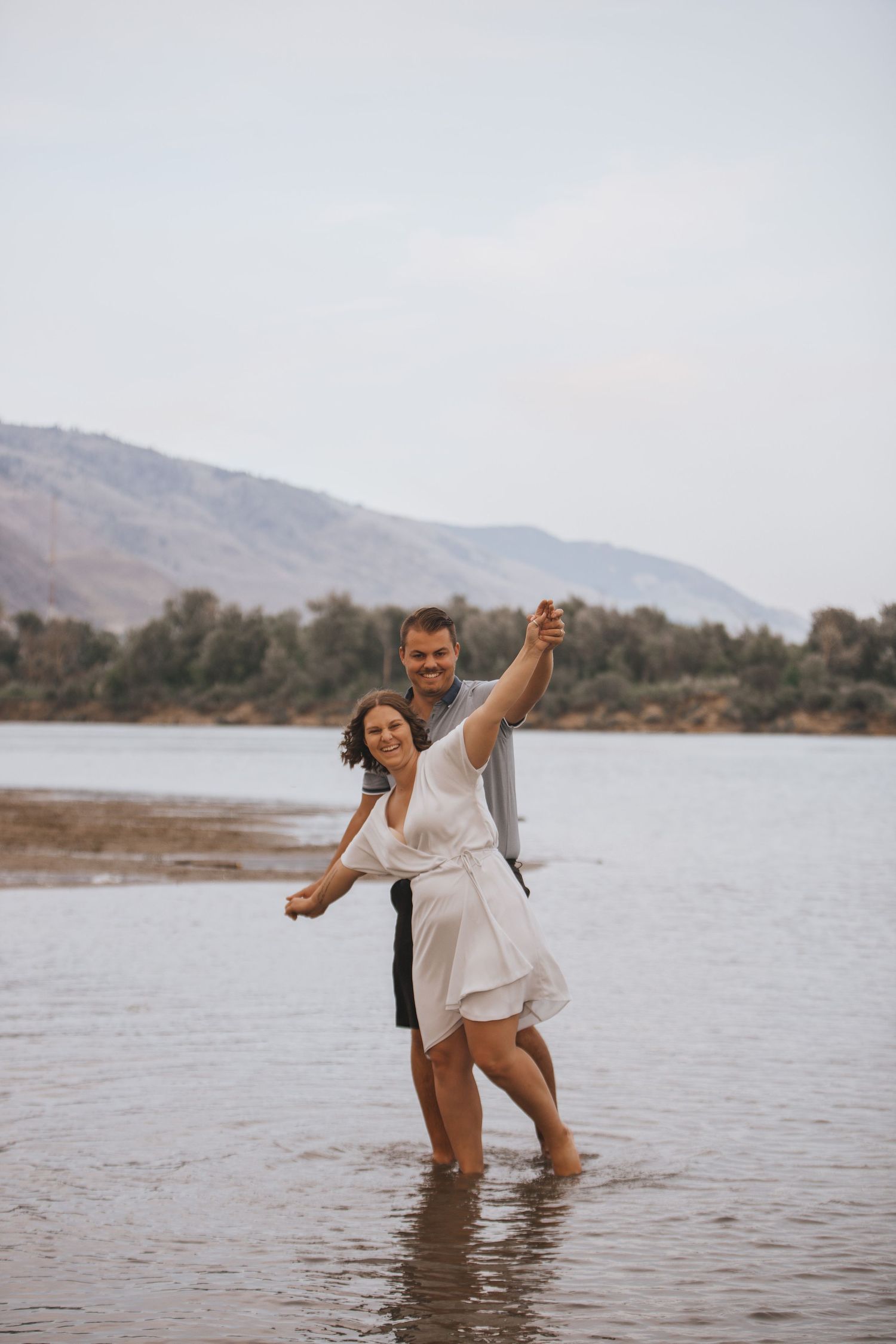 A couple splashes and dances together in shallow water with mountains in the background.