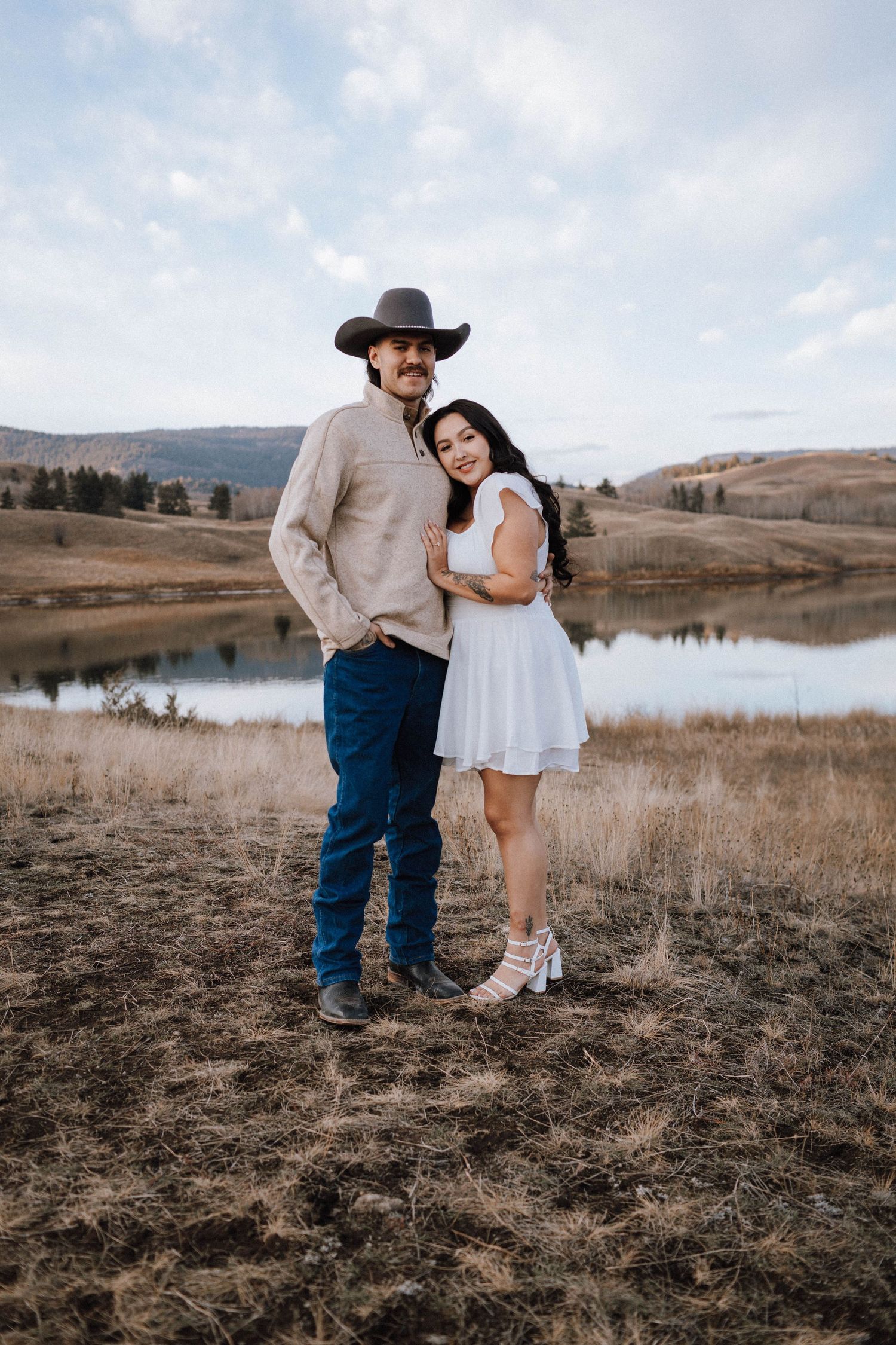 A couple poses together on a hillside overlooking a serene lake during a romantic photoshoot at sunset.