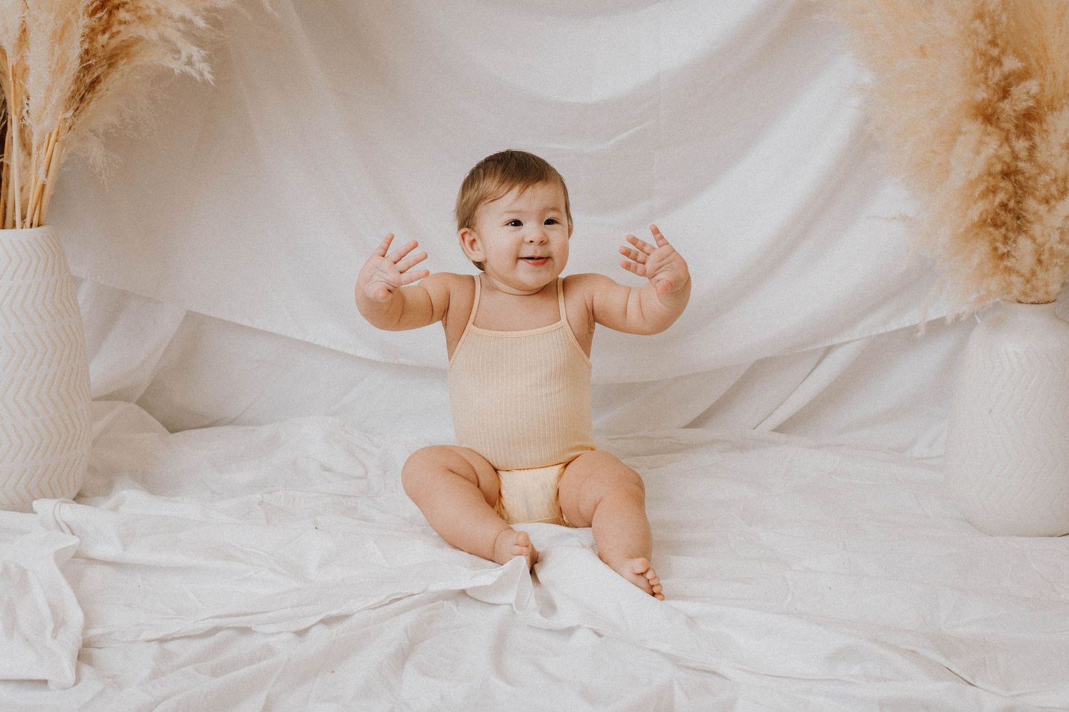 Baby wearing beige bodysuit lying on white fabric with raised arms and pampas grass decor.