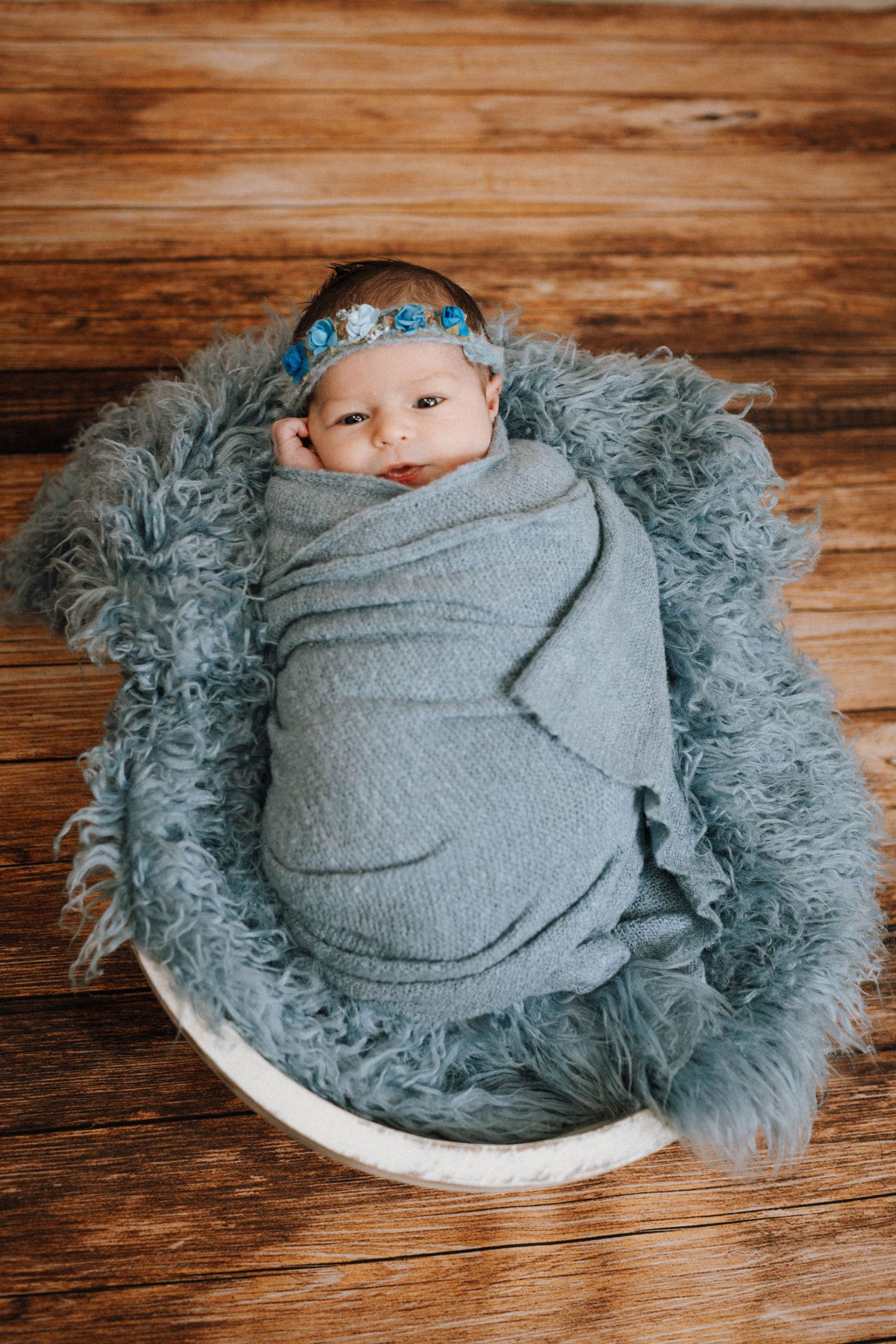 Newborn wrapped in gray swaddle with blue headband resting in furry nest against wooden background.