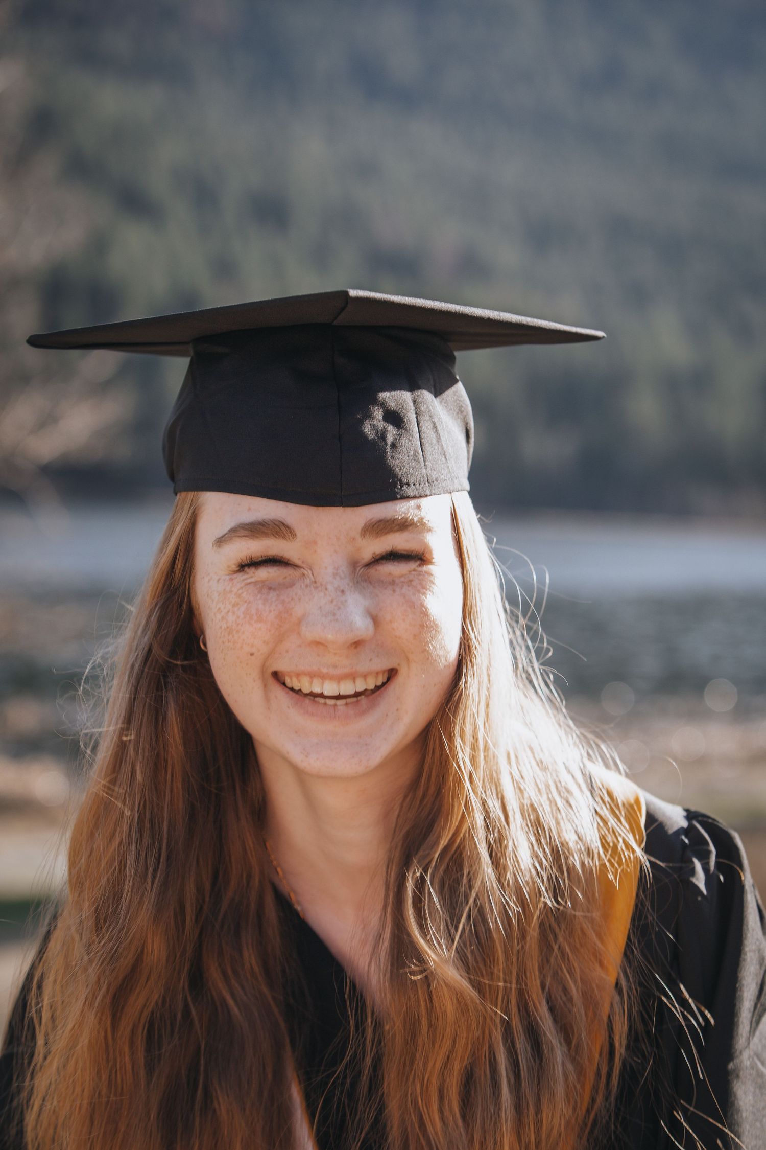Smiling graduate in cap and gown poses outdoors with mountains in background during sunny day.