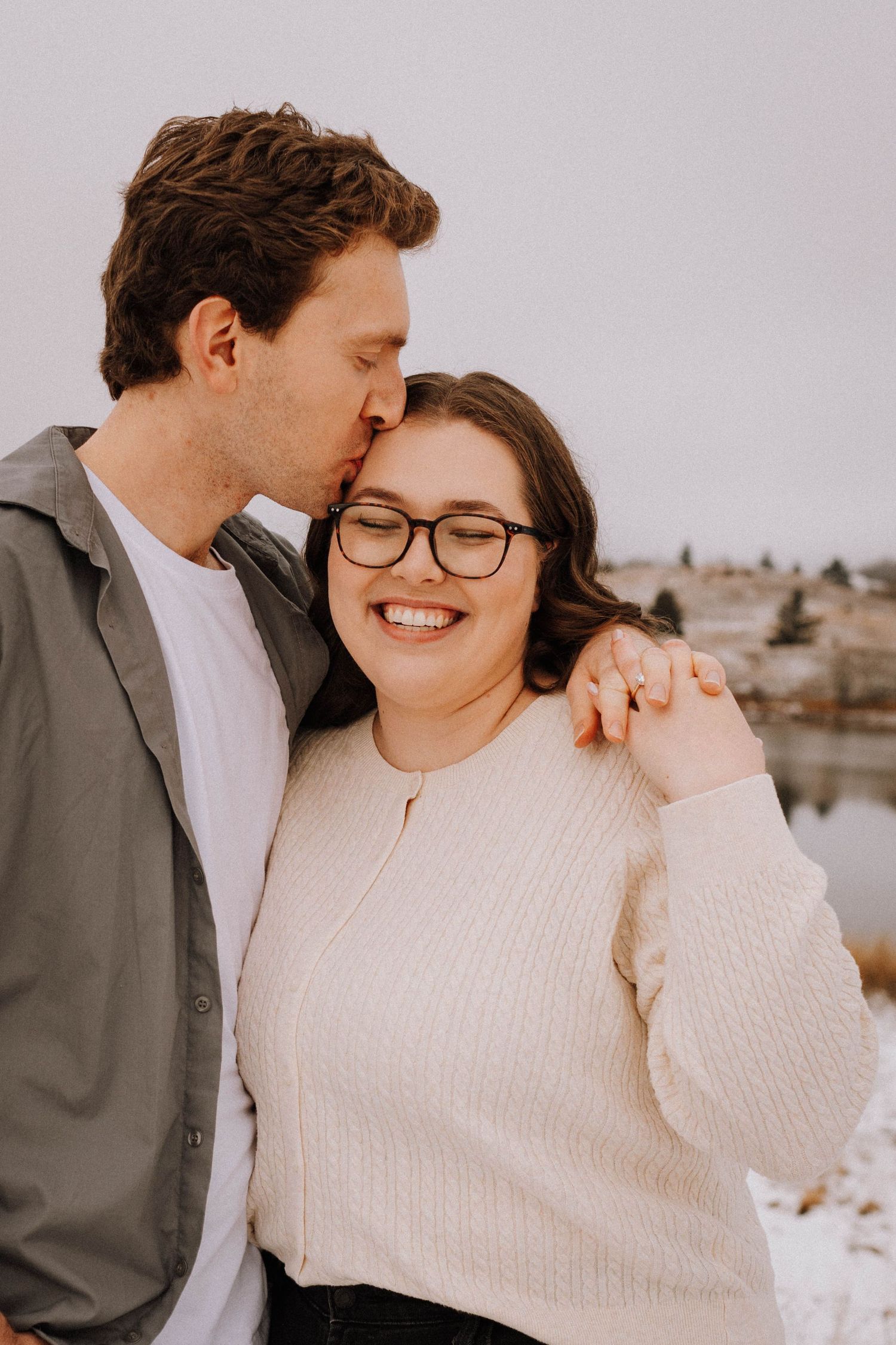 A romantic winter photo shoot showing a couple in warm sweaters sharing tender moments against a snowy landscape.