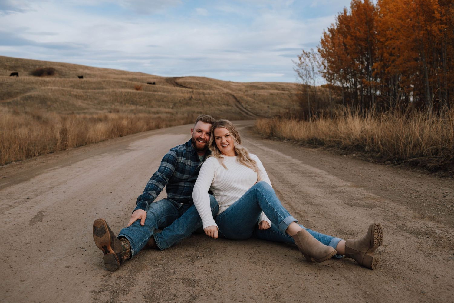Couple sits casually on a dirt road surrounded by autumn foliage and rolling hills.