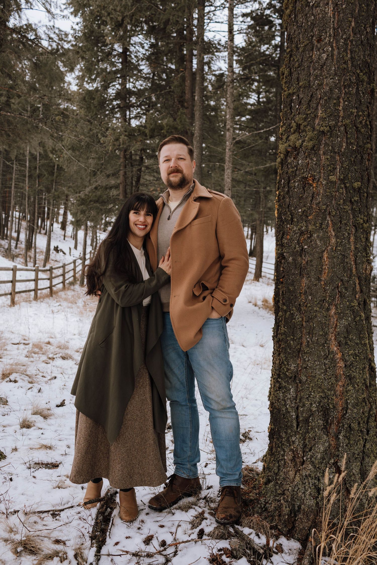 Couple in winter coats poses against snowy forest backdrop.