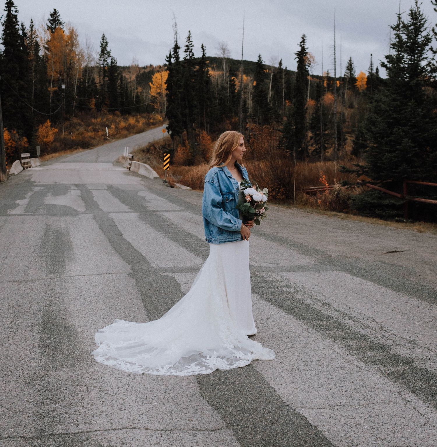 A bride in a white dress and denim jacket holds flowers on a mountain road during autumn.