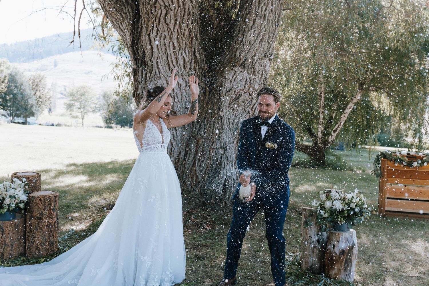 A joyful wedding moment captured under a large tree with rustic decor and falling confetti.
