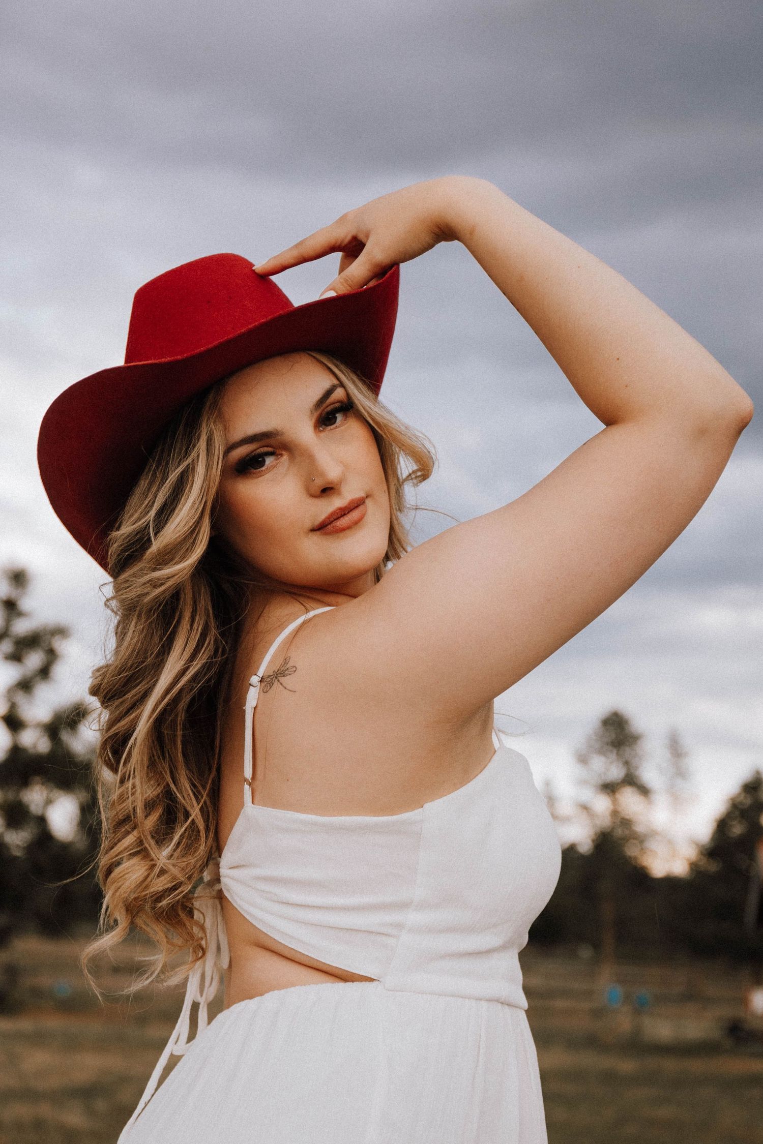 Portrait of someone in a white dress and red cowboy hat posing outdoors against a stormy sky at sunset.