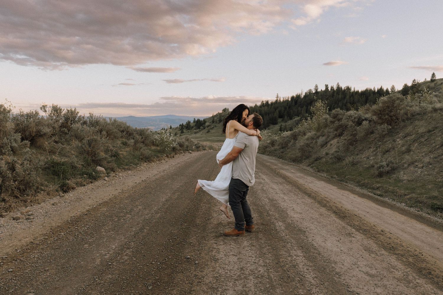 A couple embraces on a dirt mountain road at sunset with scenic hills and clouds in the background.