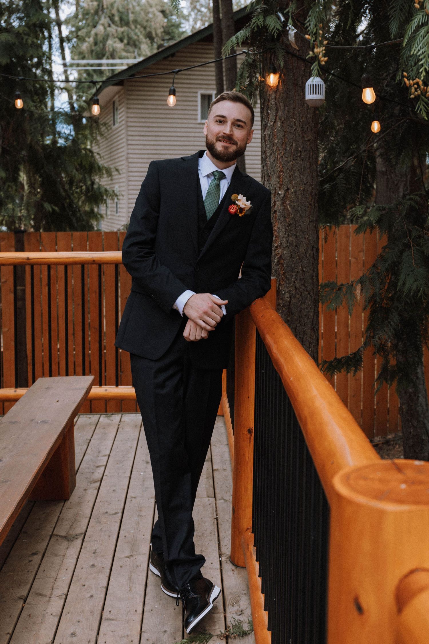 A person in a black suit poses on a wooden deck with orange railings and hanging string lights in the background.