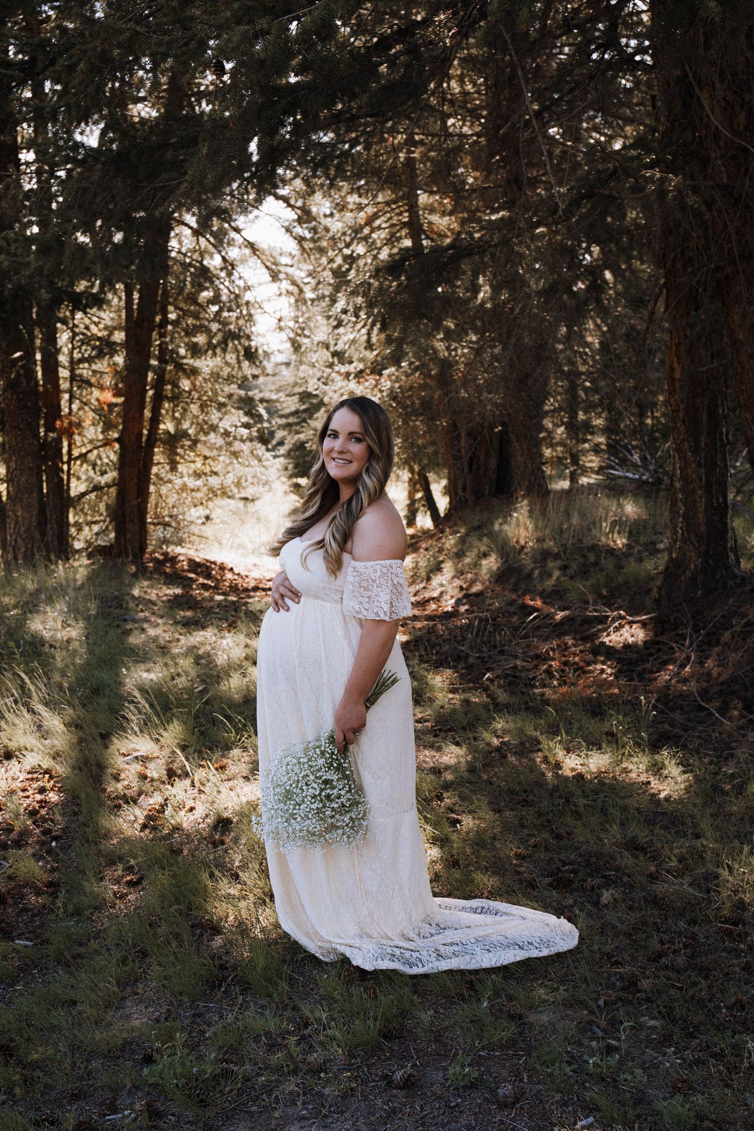 A pregnant woman in a white lace dress poses in a sunlit forest holding baby's breath flowers.