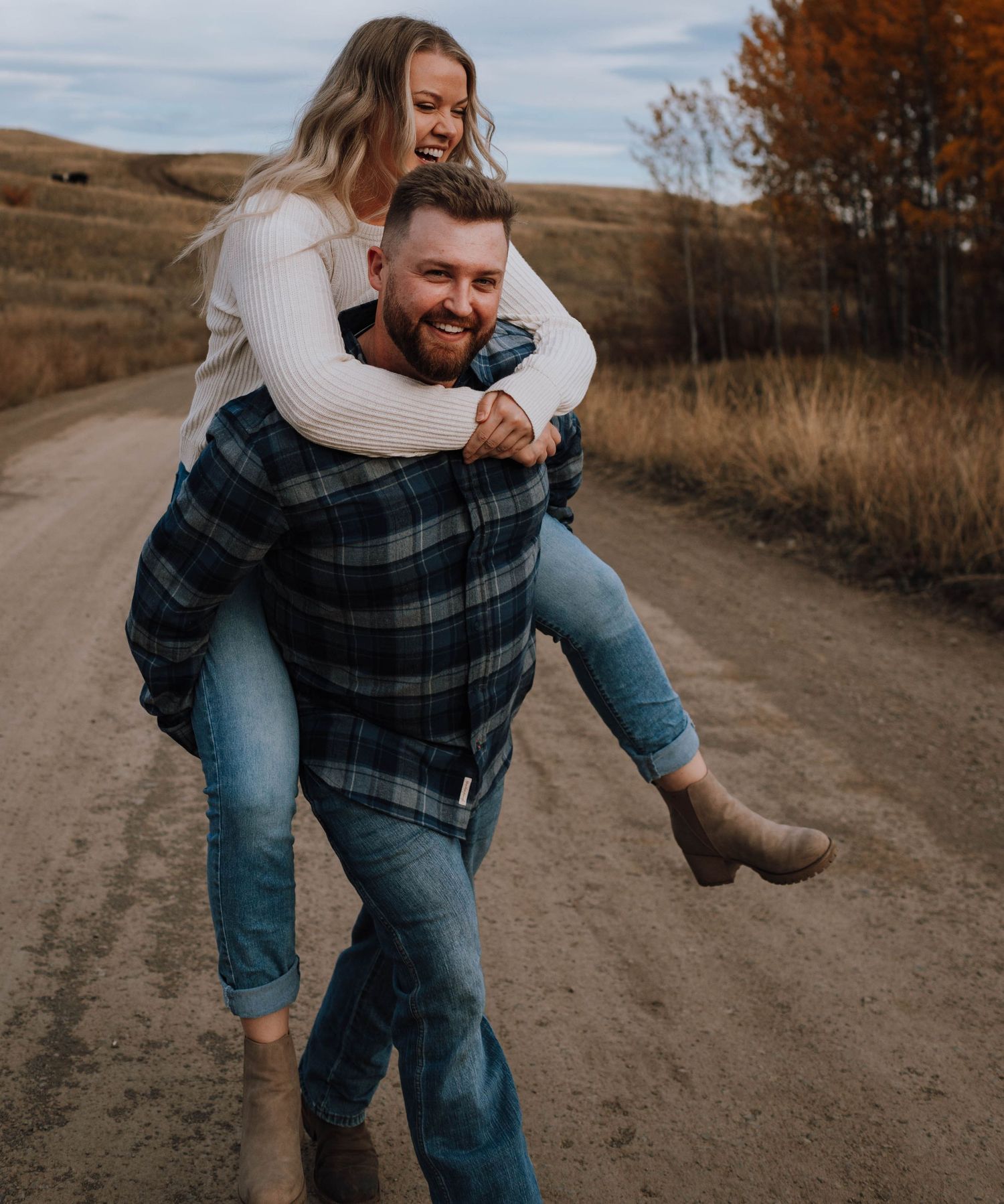 Couple shares playful piggyback ride moment on dirt road in autumn landscape.