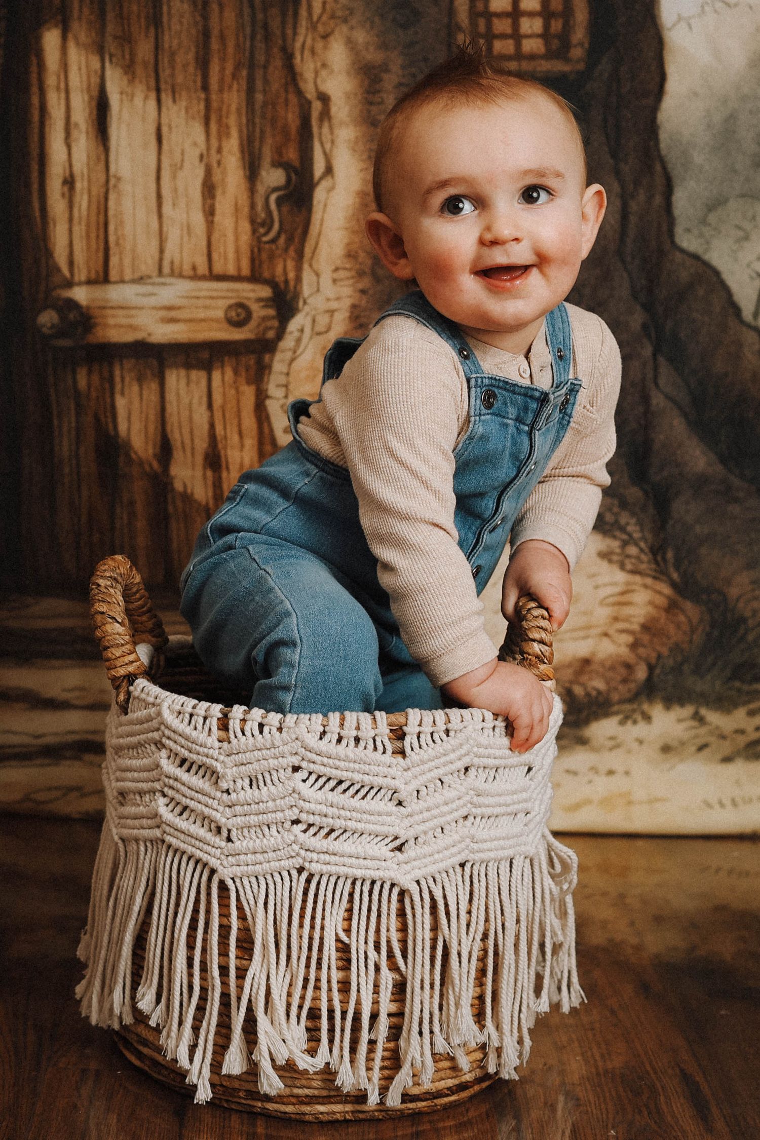 Joyful baby in overalls and cream sweater sitting on fringed macrame ottoman.