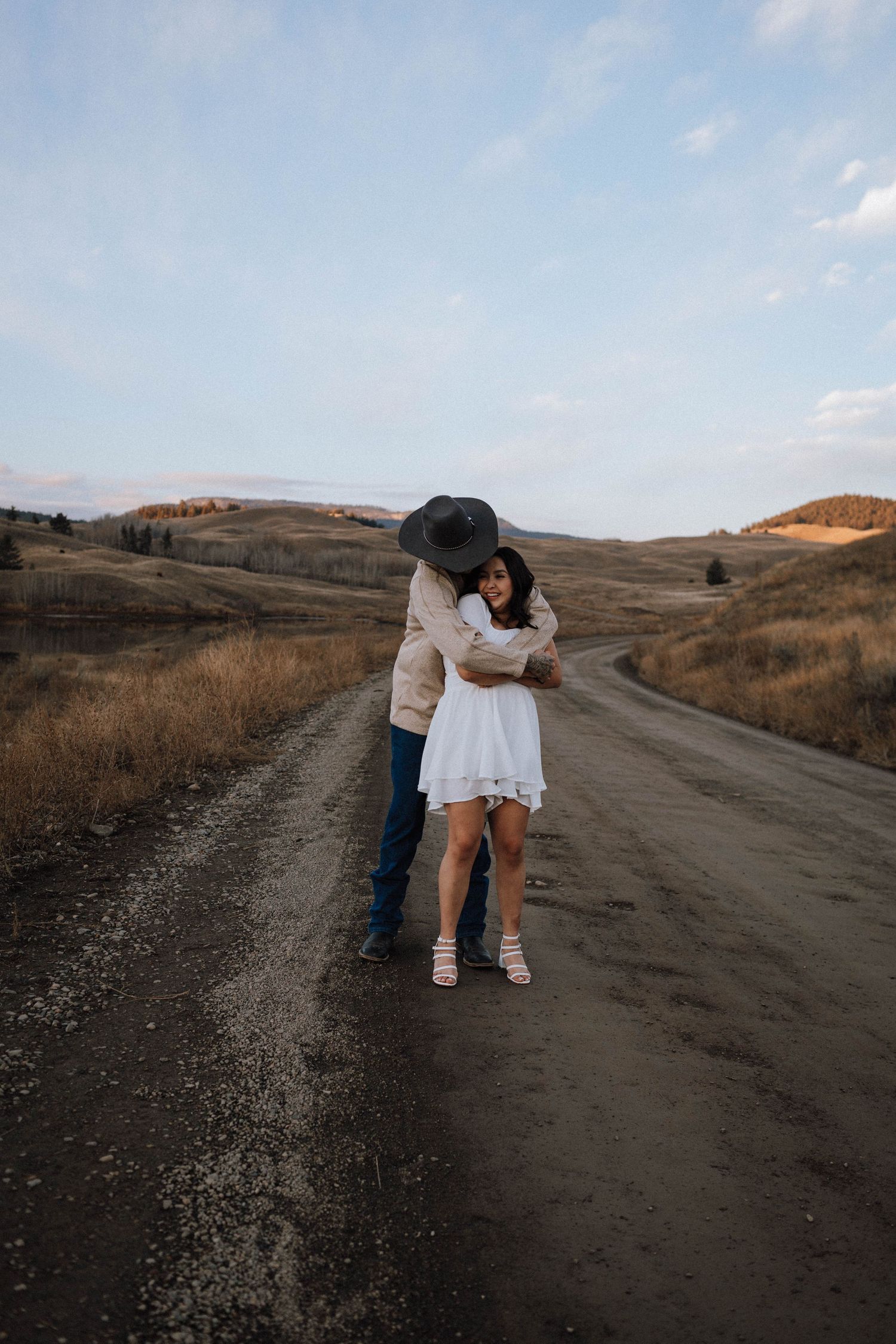A couple embraces on a dirt road through rolling hills at sunset, creating a romantic silhouette against the landscape.