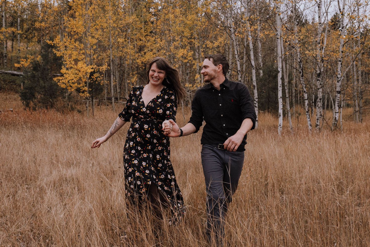 A couple walks together through a golden autumn field with tall grass and birch trees in the background.