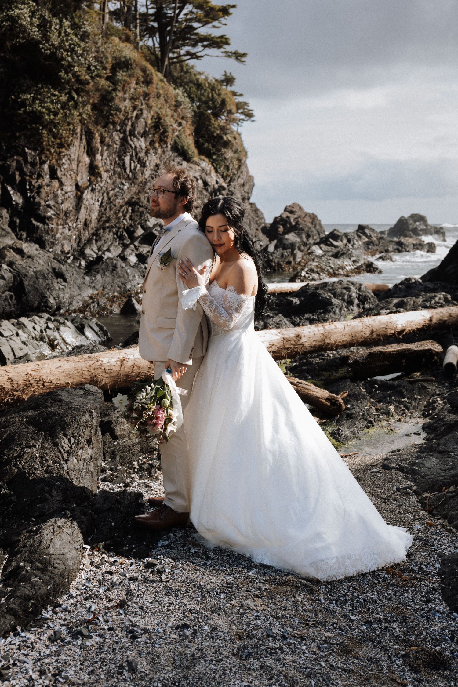 Bride and groom pose on driftwood at rocky Pacific coast beach, she in white lace gown, he in tan suit.  Ucluete Bc Black Rock Resort