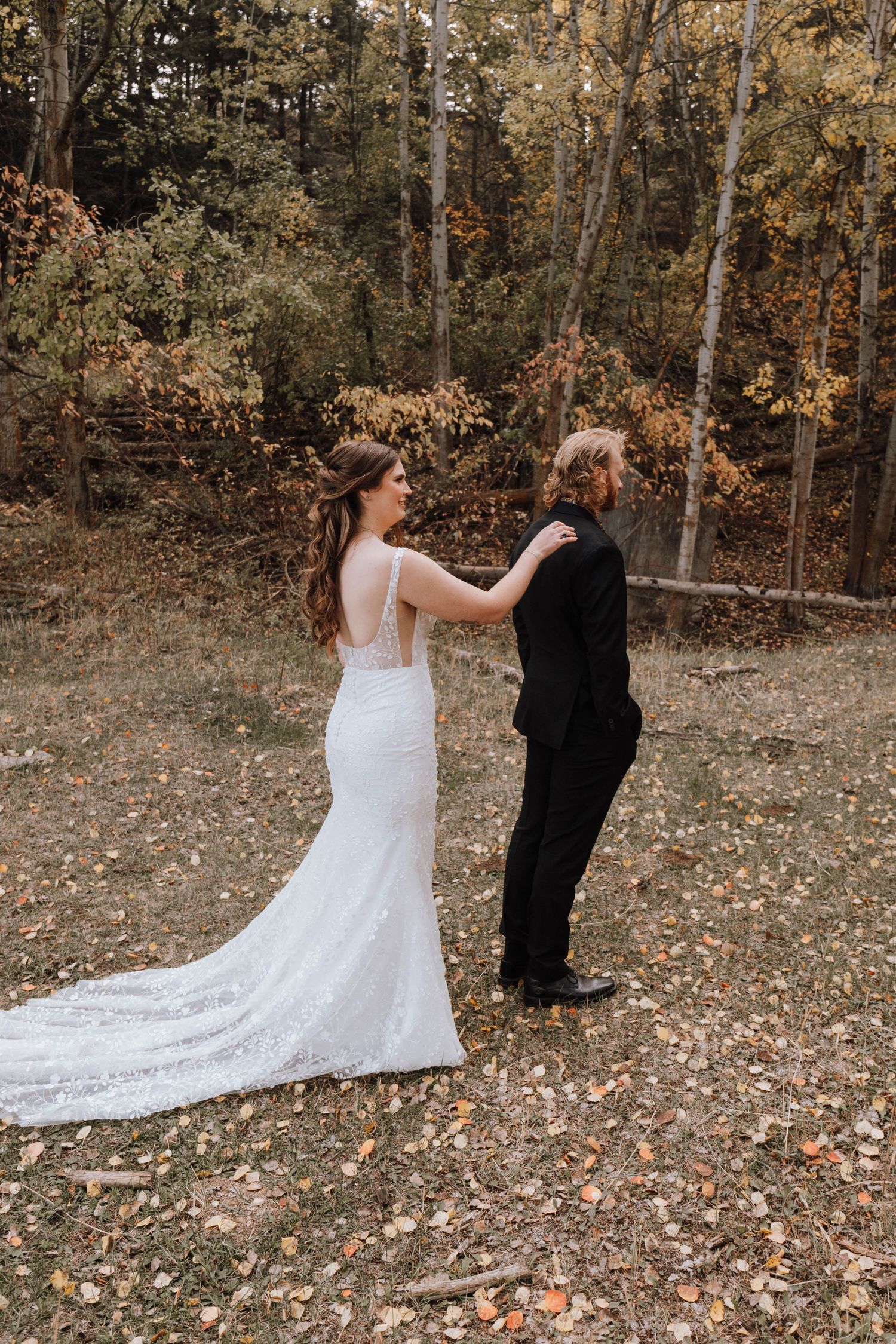 A bride touches her partner's shoulder during their first look in a wooded setting.