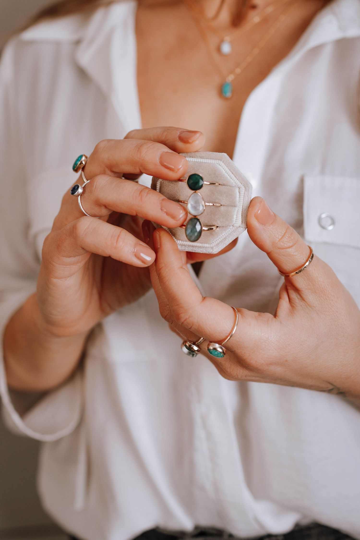 Hands wearing multiple gemstone rings holding a clear display case with jewelry.
