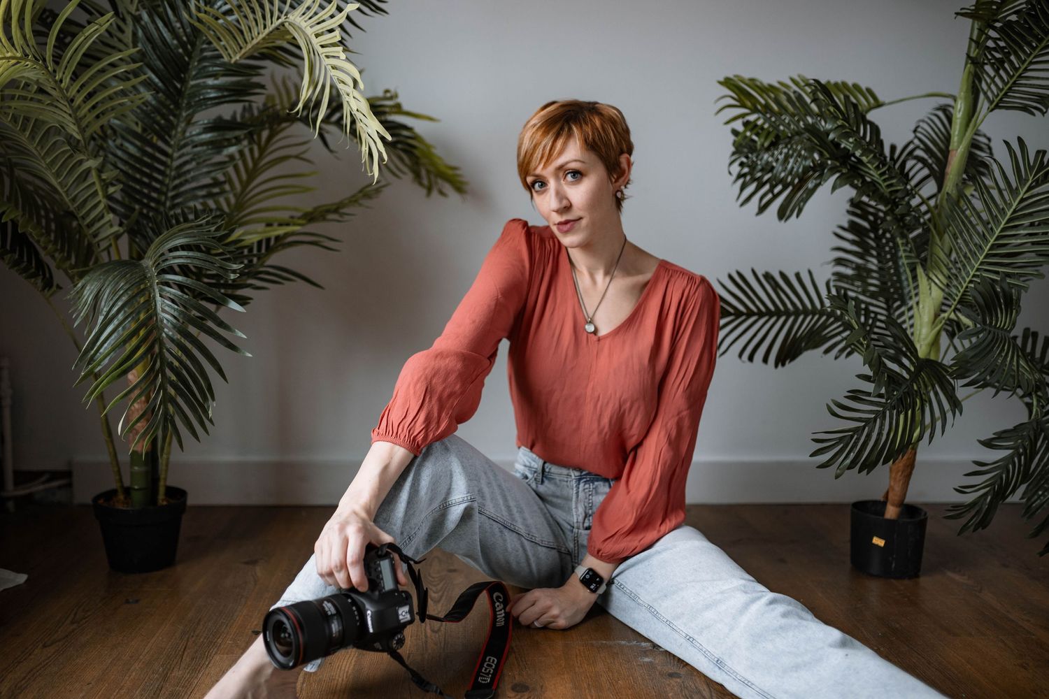 Indoor portrait session with houseplants showing photographer in coral blouse and jeans sitting on hardwood floor.