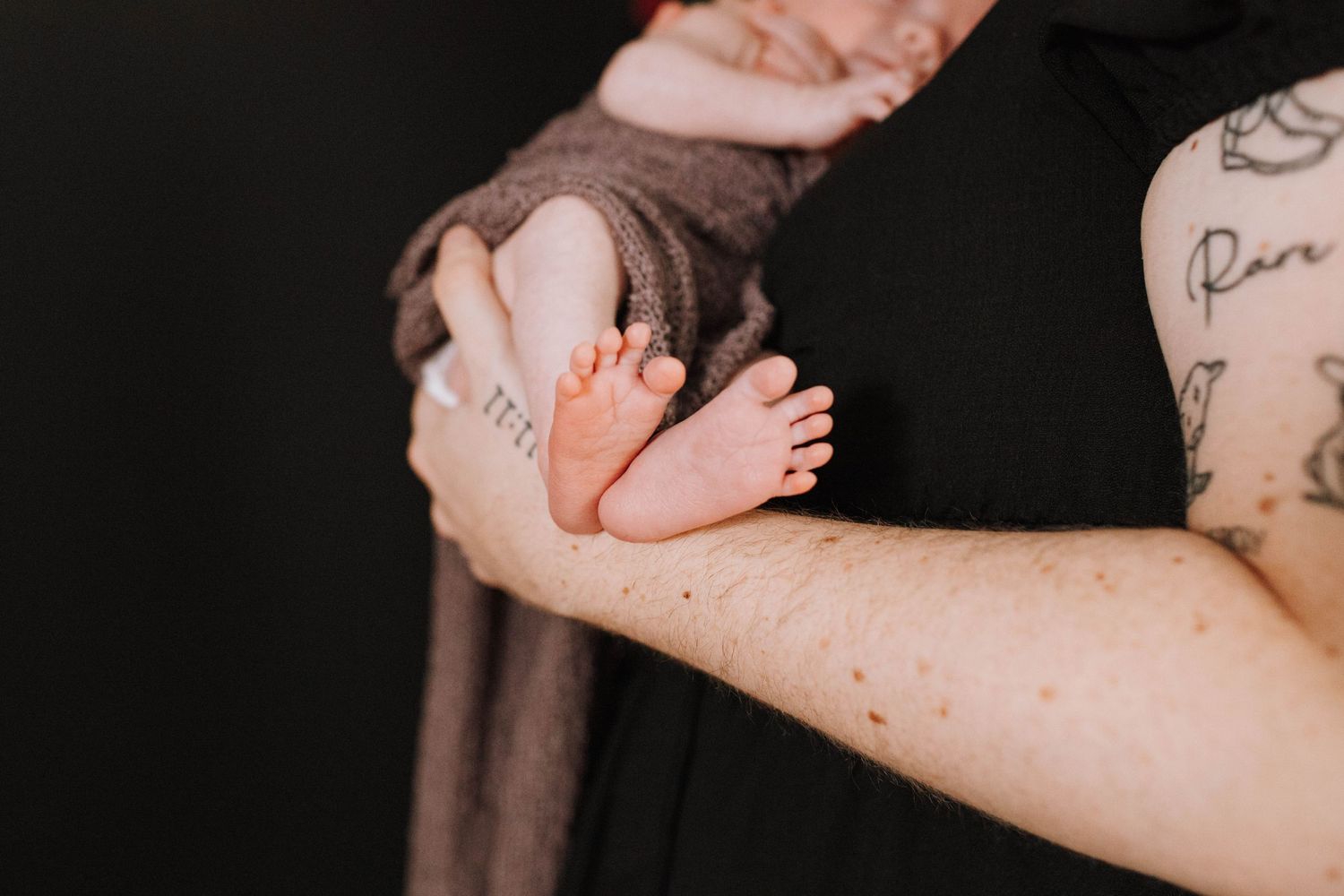 Close-up of tiny baby feet resting on person's arm with tattoos.