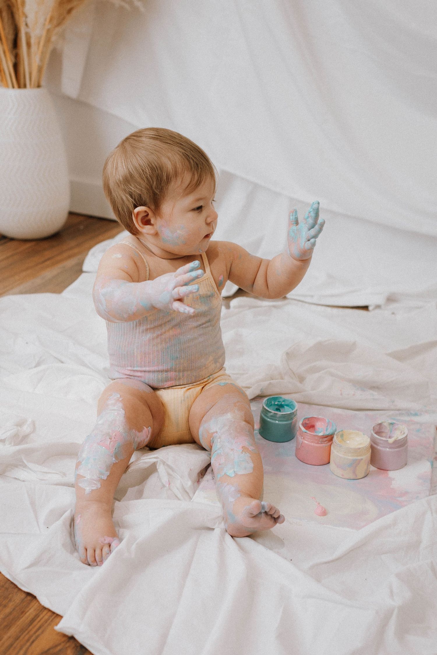 Baby playing with paint while sitting on white sheet with pampas grass decor.