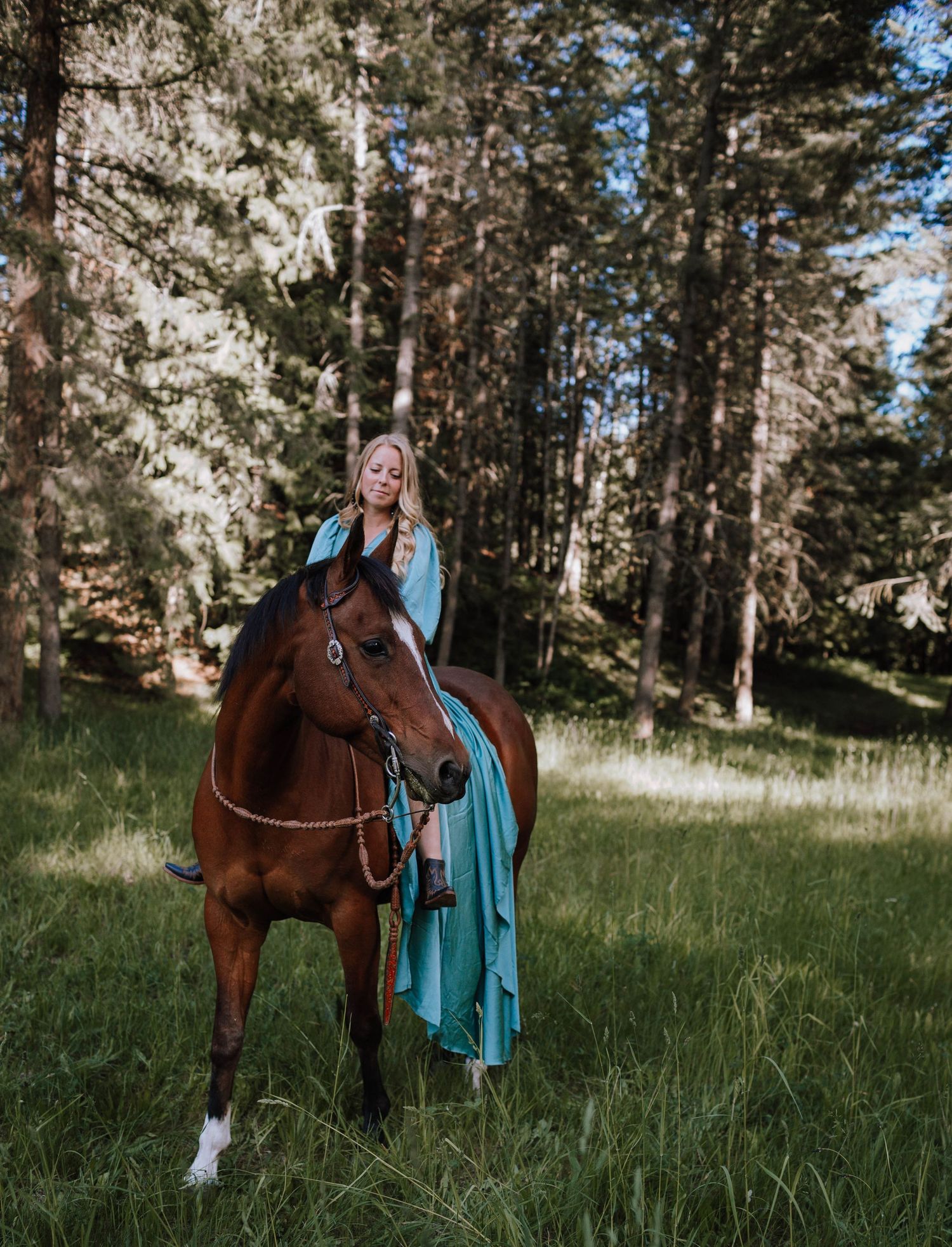 Equestrian in flowing turquoise dress sits atop brown horse in forest clearing.