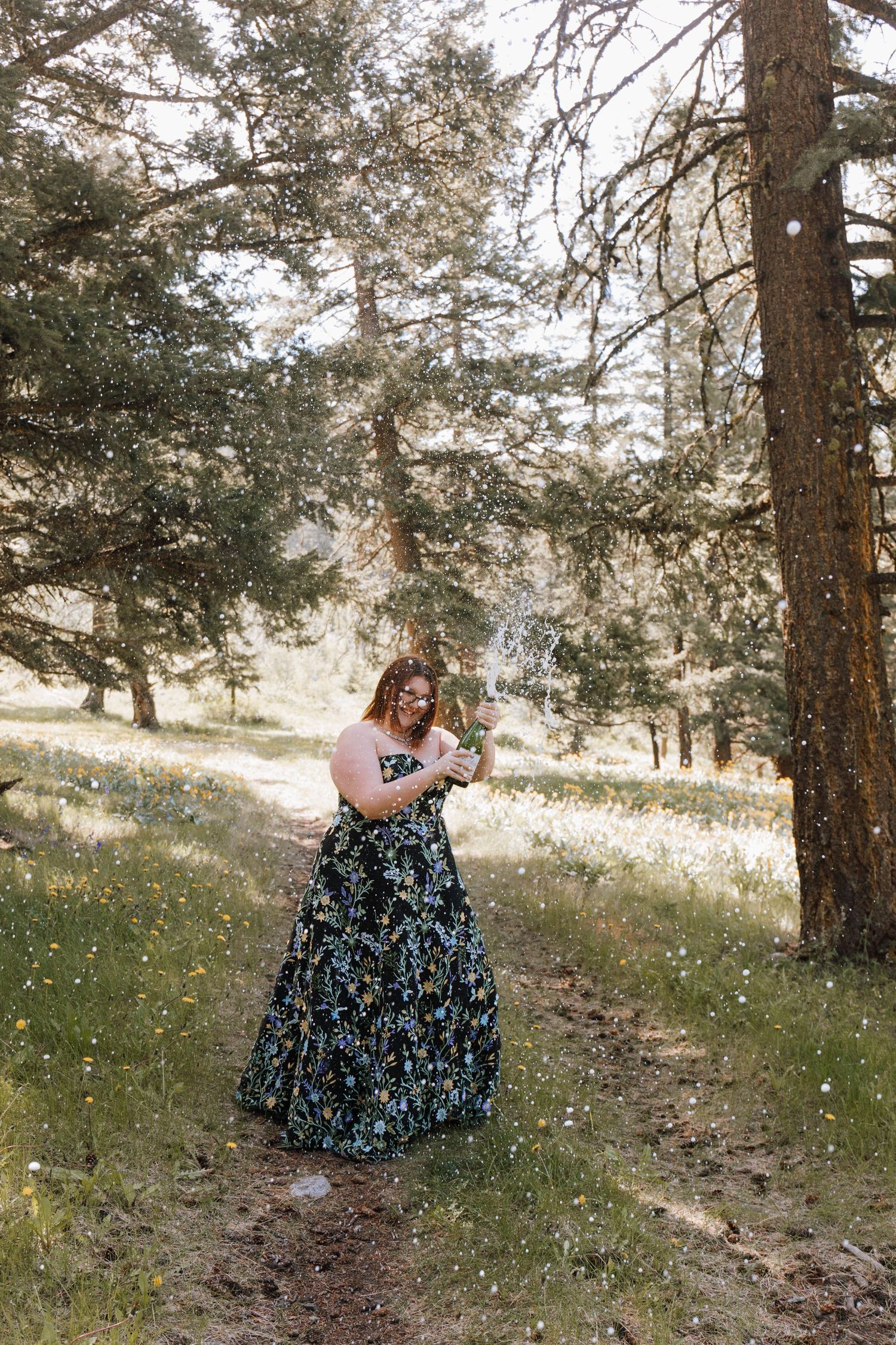 Person in a floral maxi dress stands on a forest path surrounded by sunlit pine trees and wildflowers.