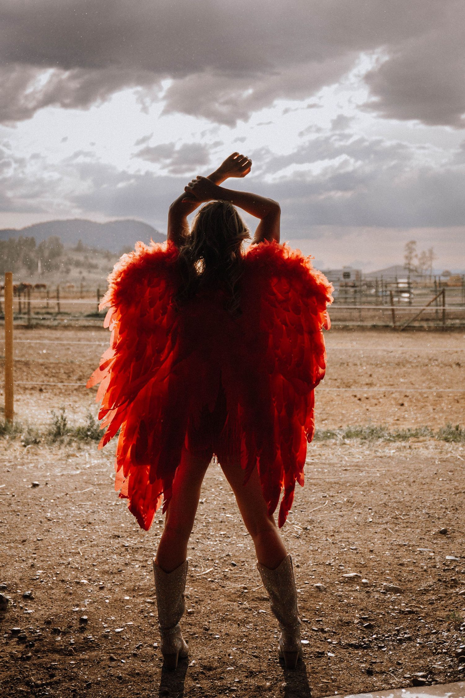 A figure wearing dramatic red feathered wings poses against cloudy skies in a rustic desert landscape.