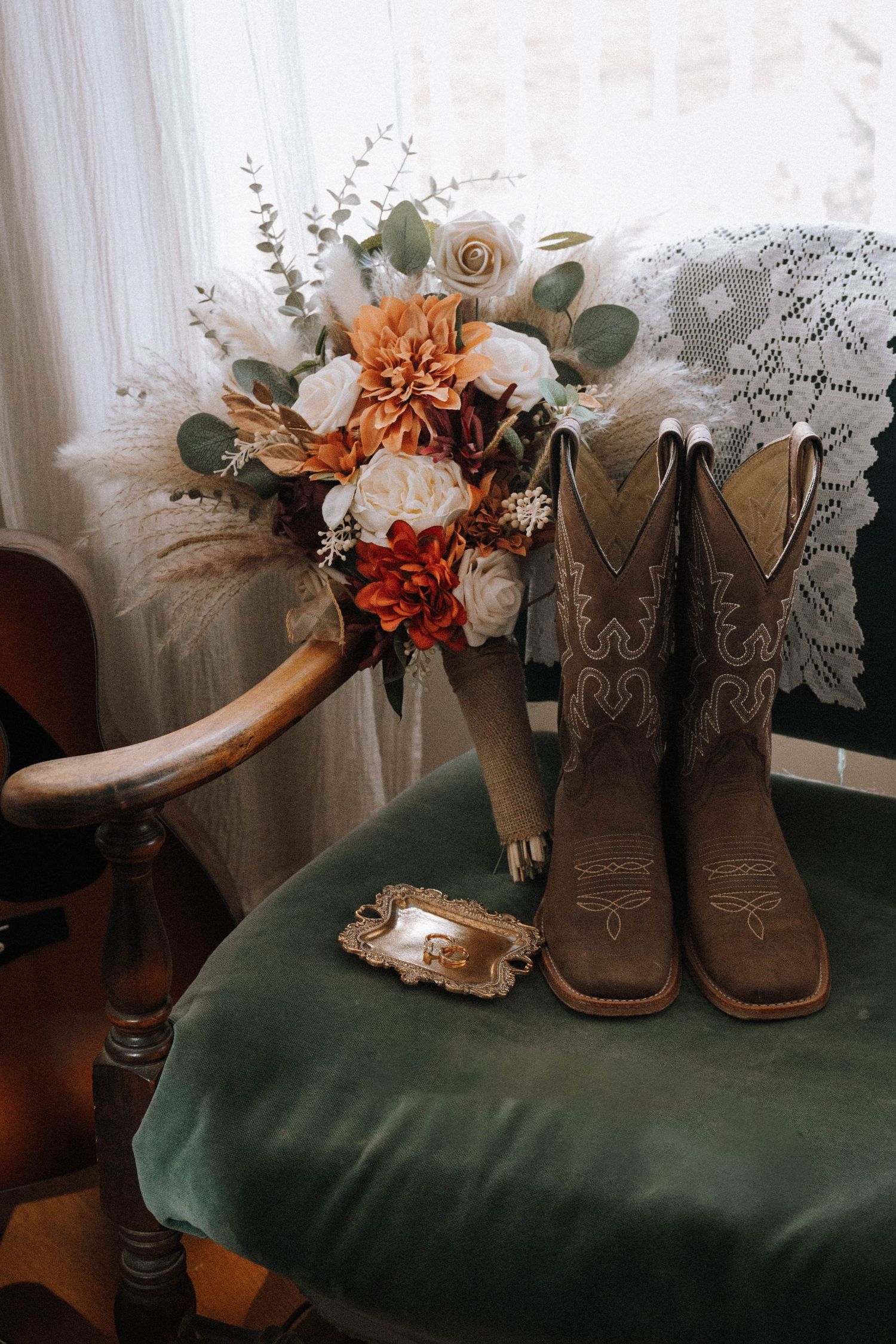 Brown leather cowboy boots decorated with orange and white flowers sit on a dark green velvet surface near lace curtains.