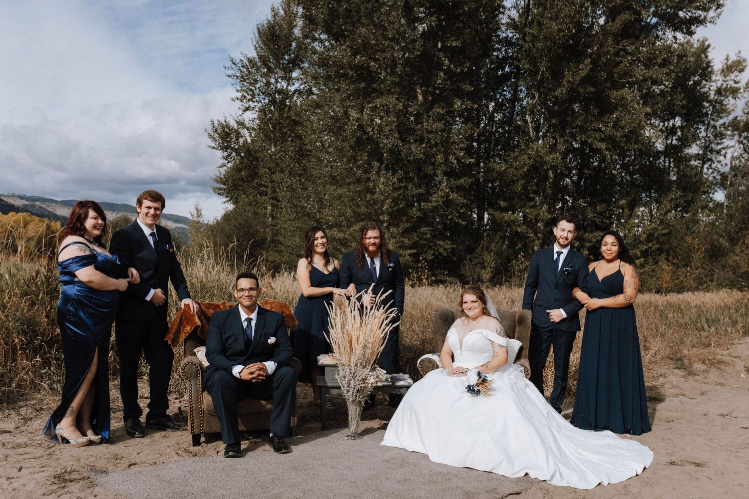 Wedding party poses together on sandy beach with evergreen trees in background during outdoor celebration.