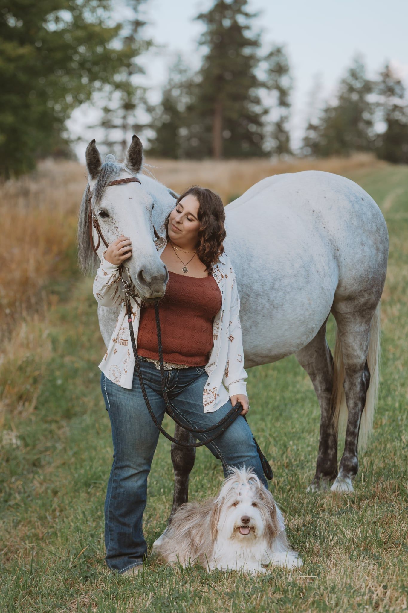A person stands with a white horse and fluffy white dog in a grassy field at dusk.