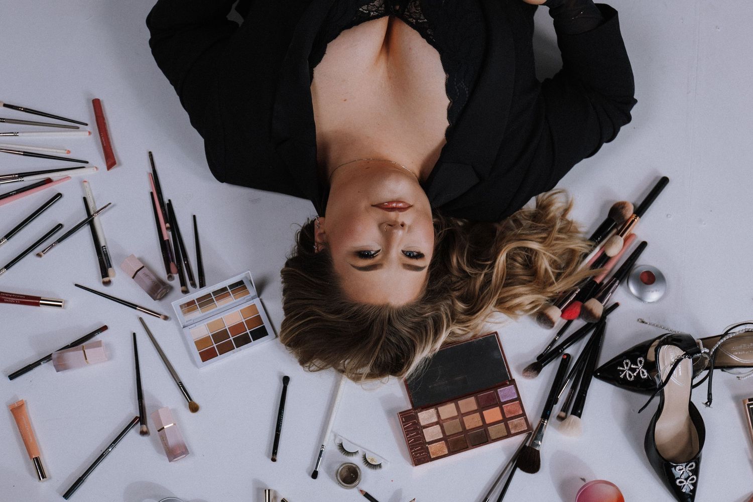 Beauty products and makeup brushes scattered on white surface with hands reaching for cosmetics.
