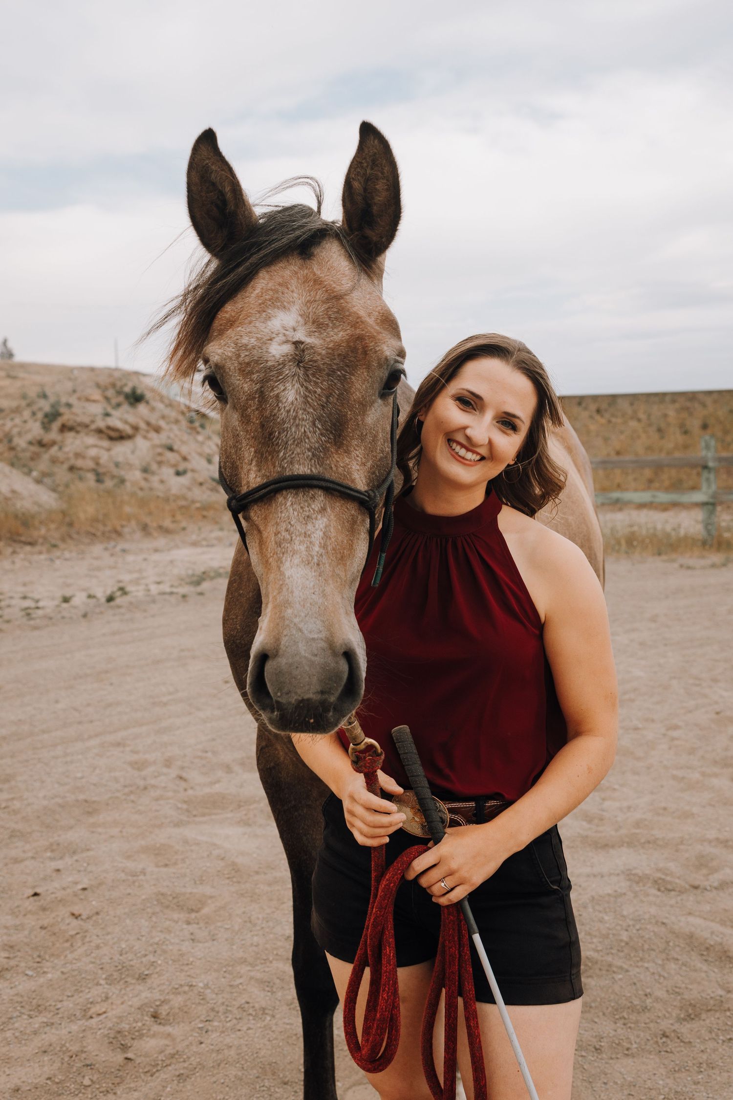 Smiling young woman in burgundy top holding red lead rope beside a brown horse in sandy outdoor arena.