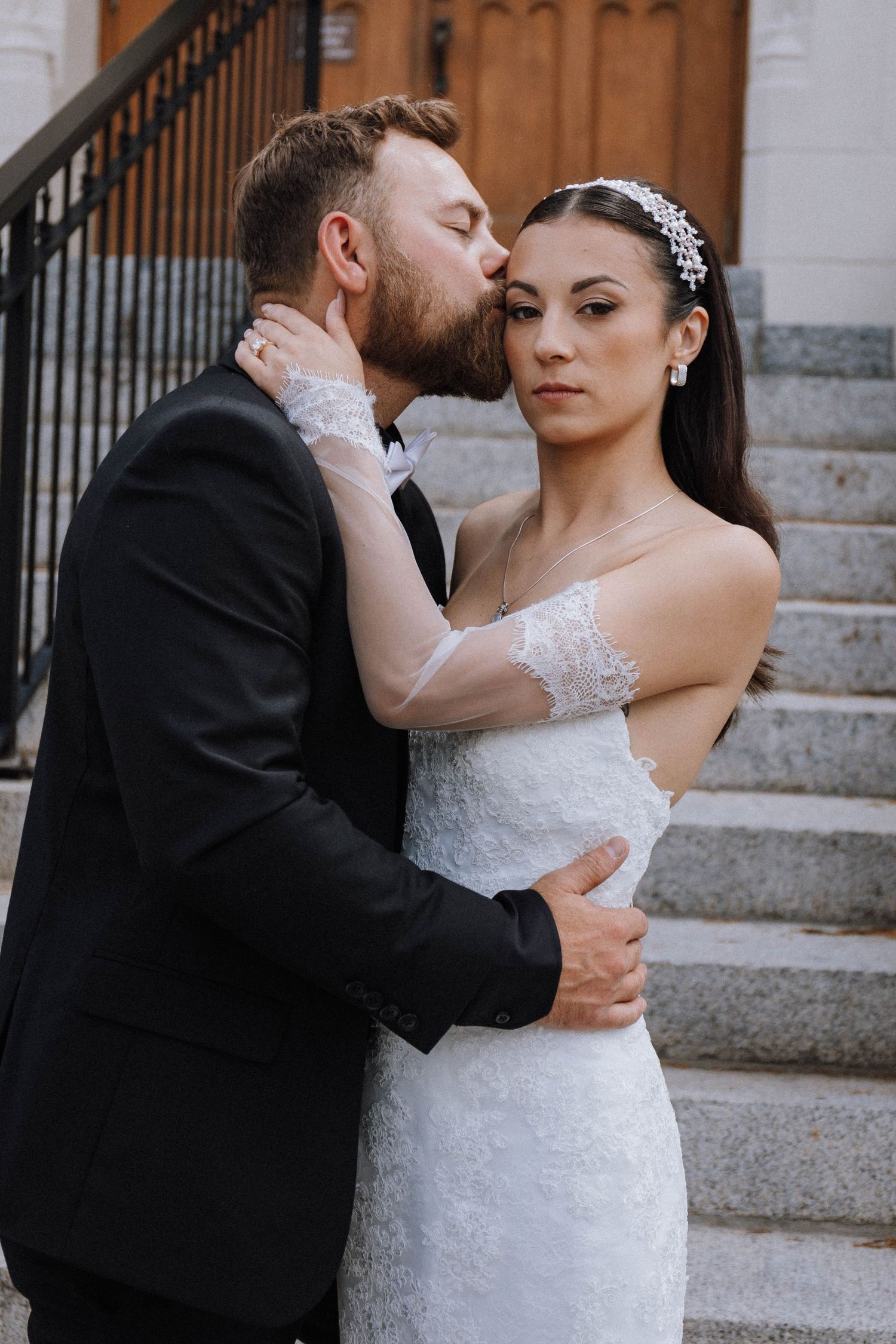 Romantic wedding couple embraces on stone steps, bride in lace gown with crystal headpiece and groom in classic tuxedo.