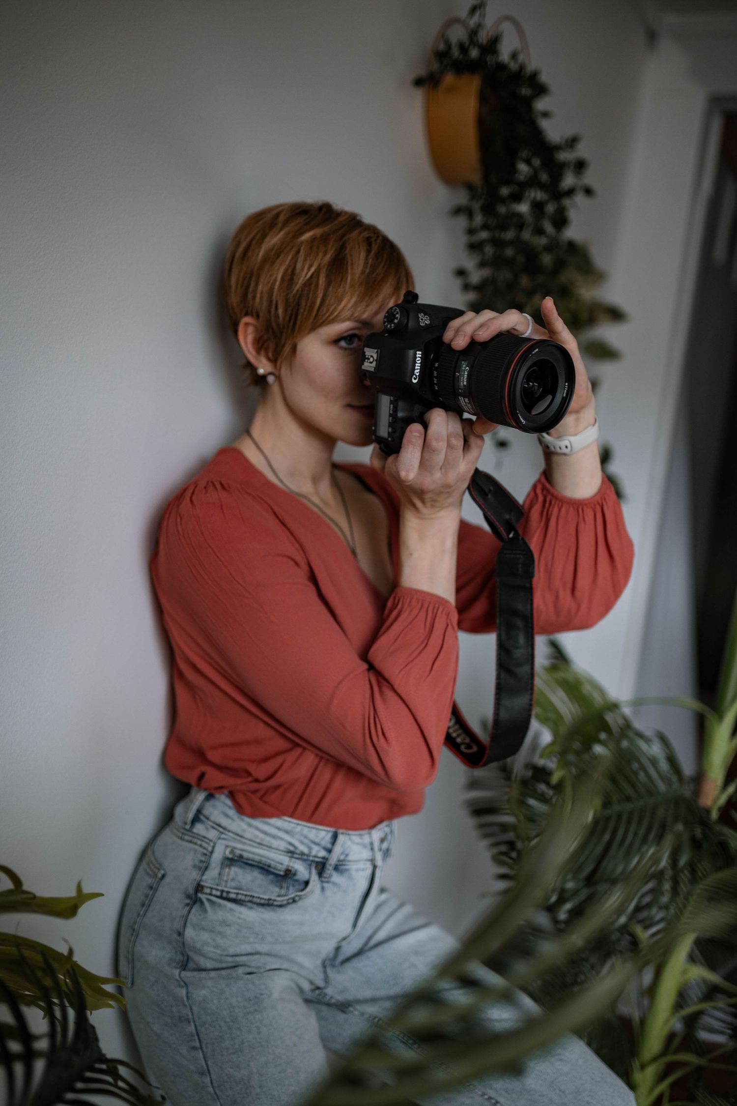 Photographer in coral blouse taking pictures with professional camera among indoor plants.