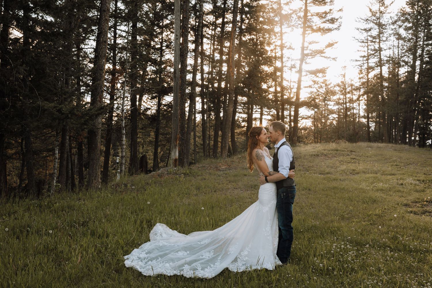 Bride and groom embrace in a sunlit forest meadow, her white gown trailing on lush grass at golden hour.