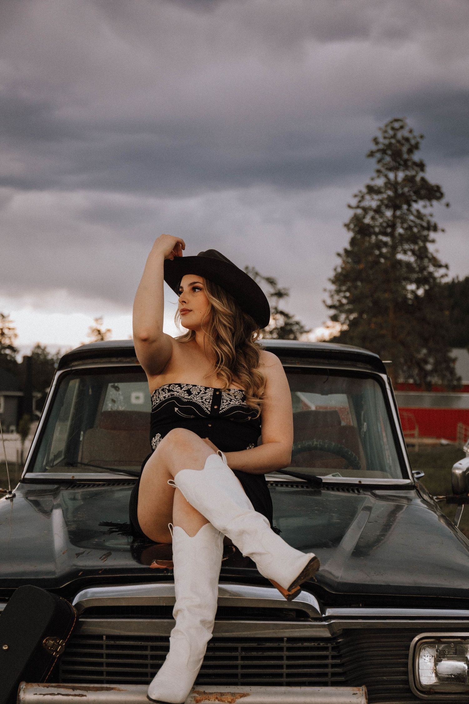 Woman in white dress poses near vintage black car against cloudy sky in moody outdoor photoshoot.