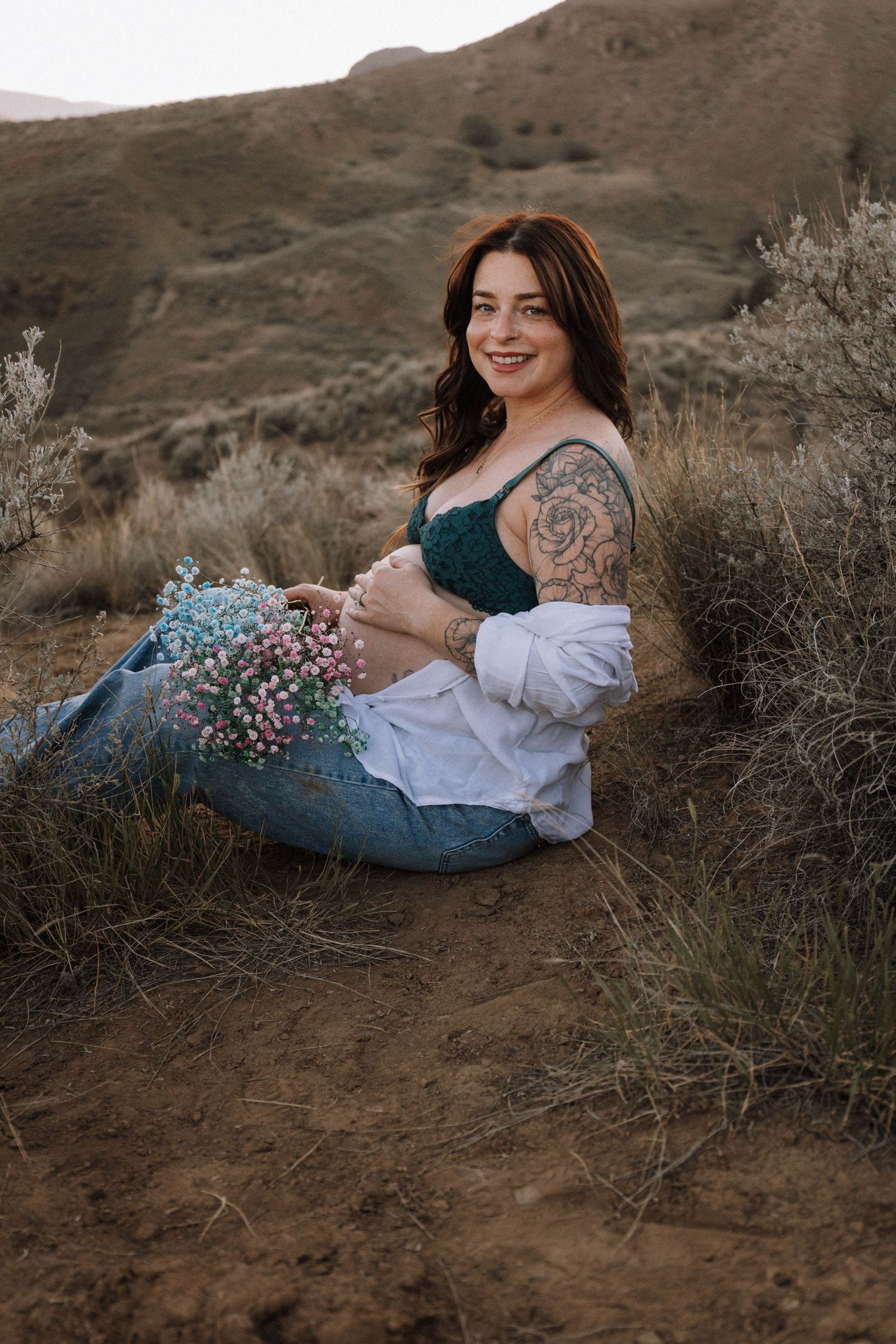A woman in a dark teal dress sits on a hillside holding wildflowers at sunset with mountains in the background.