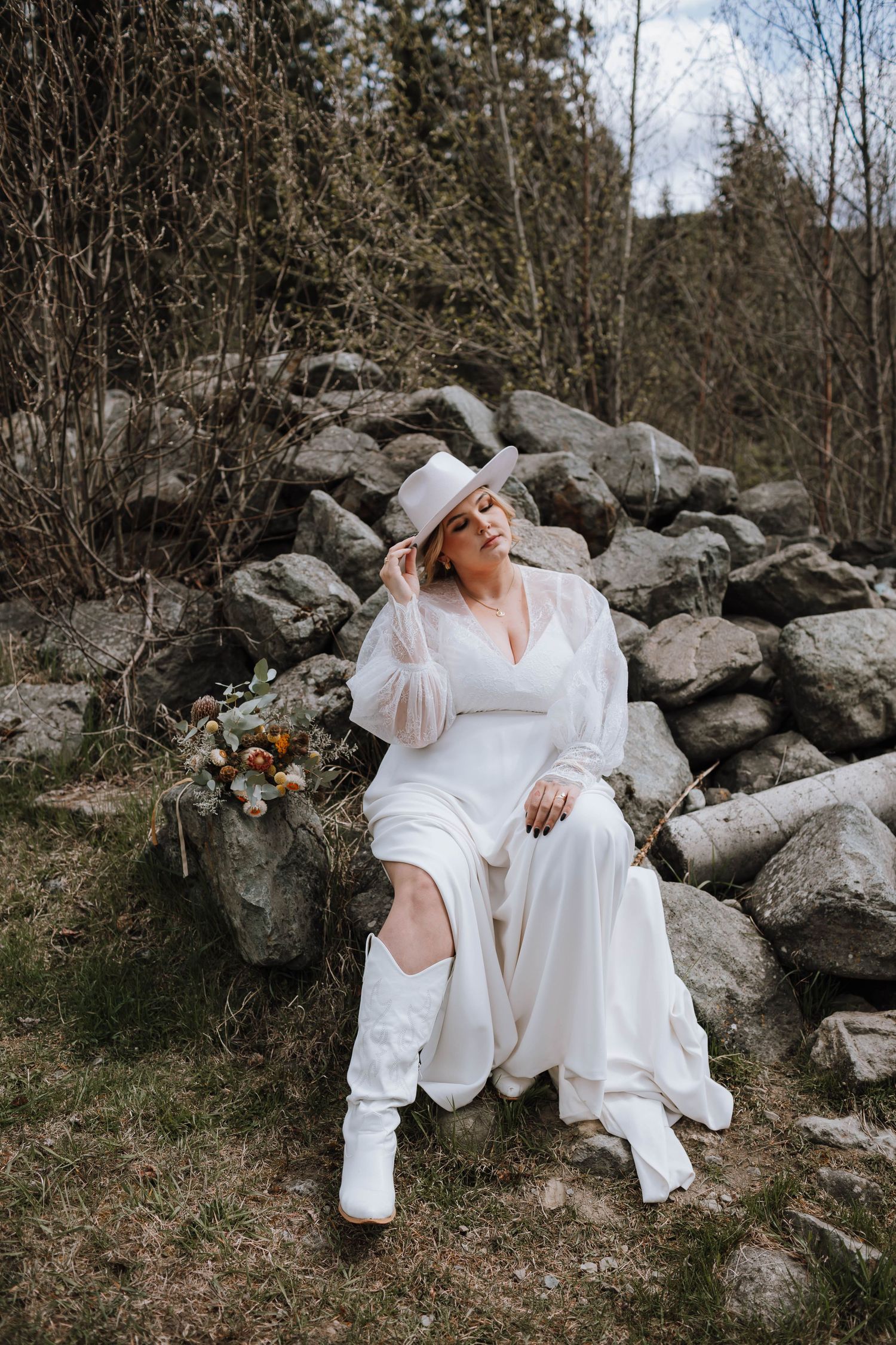 A bride in white dress and boots poses on rocks in a natural outdoor setting.