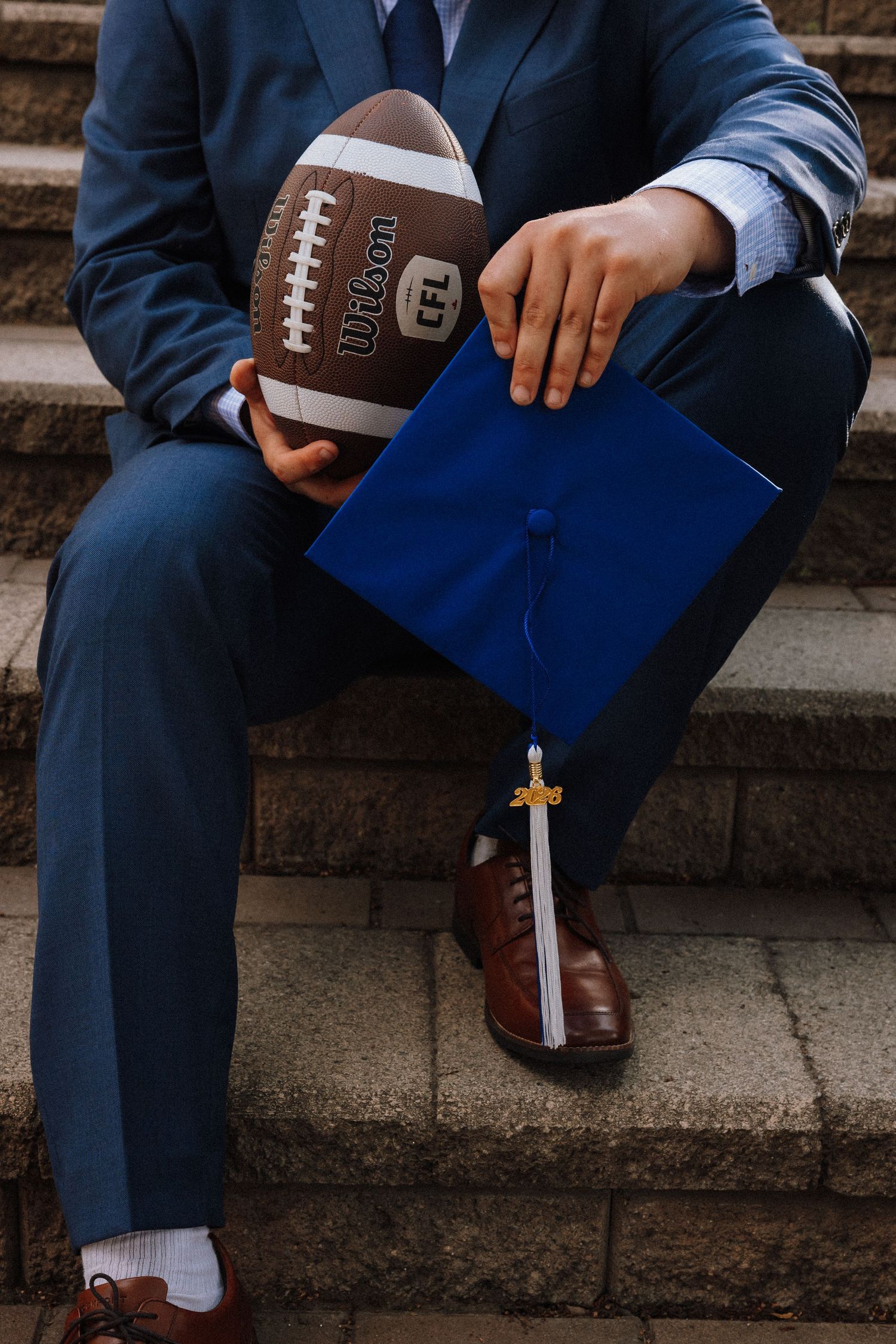 Graduate in blue suit holding Wilson CFL football and blue graduation cap while sitting on stone steps.